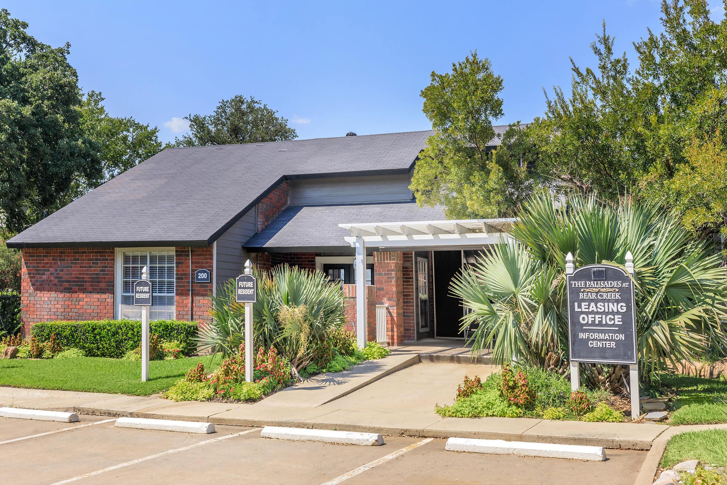 A brick building with a gray roof, featuring a leasing office sign and landscaping with palm trees and green bushes. The entrance has a covered walkway, and the parking lot is visible. The sky is clear and blue, indicating a sunny day.