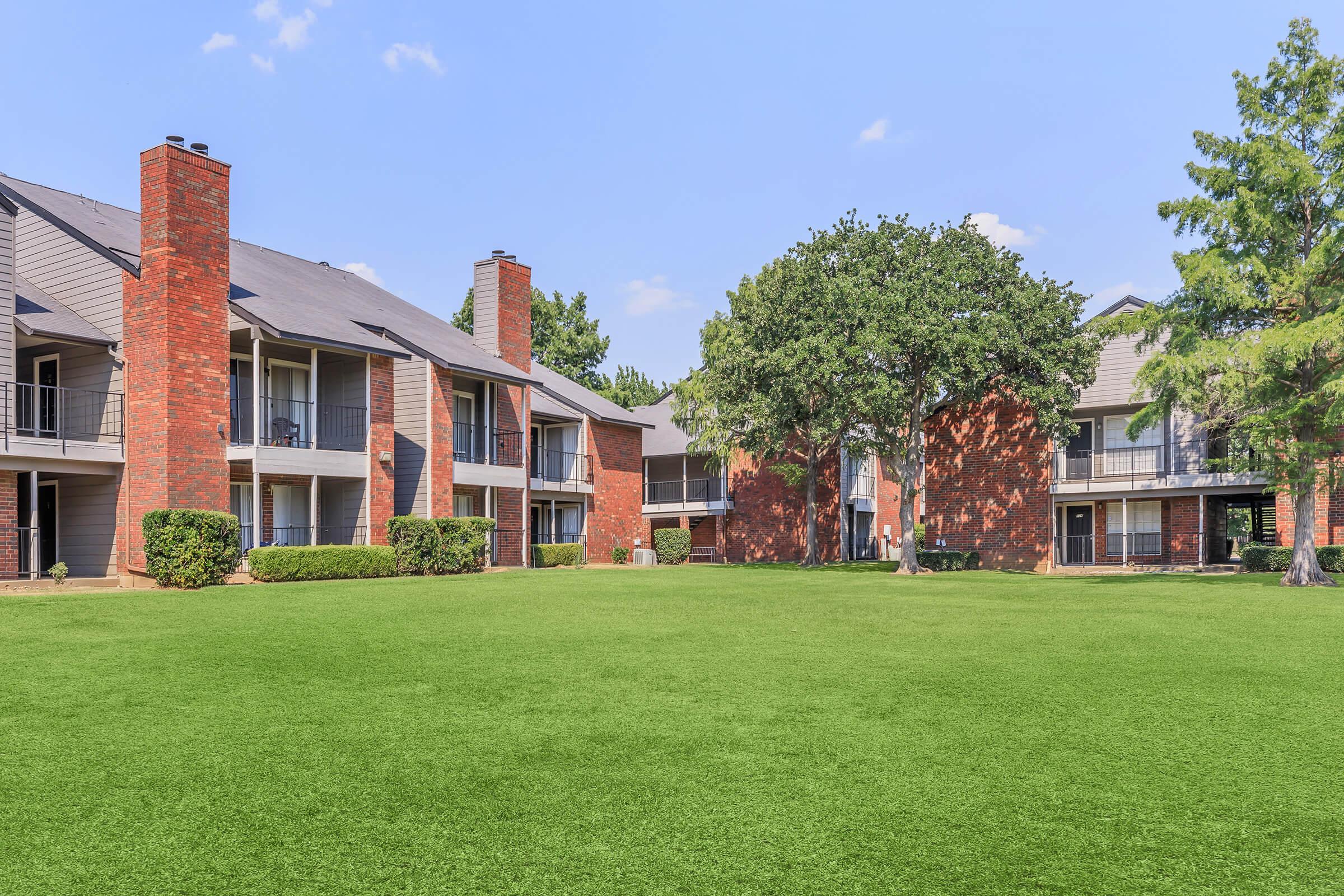 A well-maintained apartment complex featuring two-story buildings with brick exteriors. The lawns are lush and green, with trees providing shade. Clear blue sky and a serene atmosphere create a pleasant residential environment.
