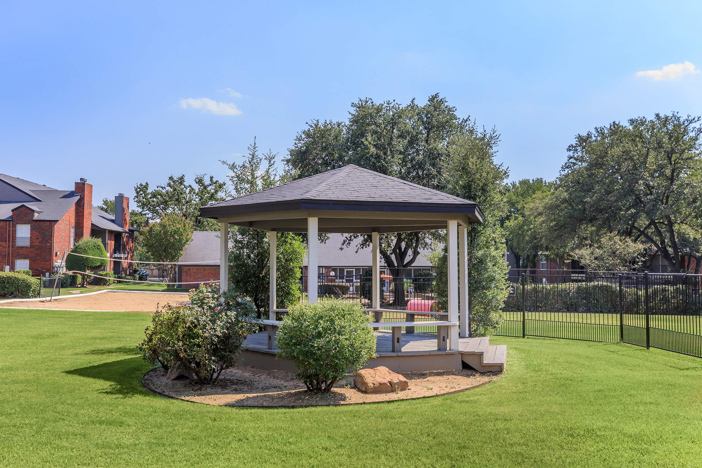 A scenic view of a gazebo surrounded by lush green grass and shrubs, with sunny skies above. The gazebo features a gray roof and white wooden structure, set in a residential area with trees in the background. A fenced area is visible, adding to the tranquil atmosphere of the outdoor space.