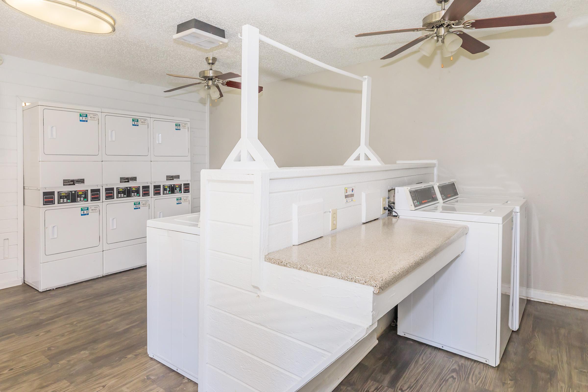 A laundry room featuring several washing machines and dryers neatly arranged against the wall, with countertops for folding laundry. The space is well-lit with ceiling fans and a clean, modern design, highlighted by a light color scheme and wooden flooring.