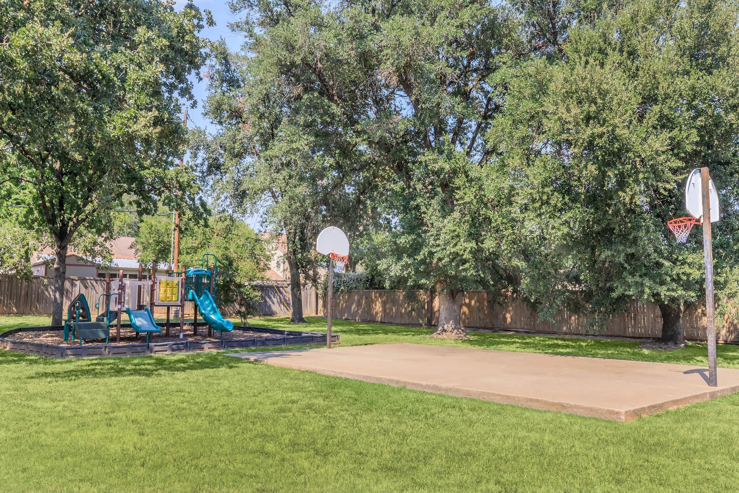 A sunny playground featuring a blue slide and climbing structure, adjacent to a basketball court with two hoops. The area is surrounded by tall green trees and a wooden fence, with well-maintained grass covering the ground.