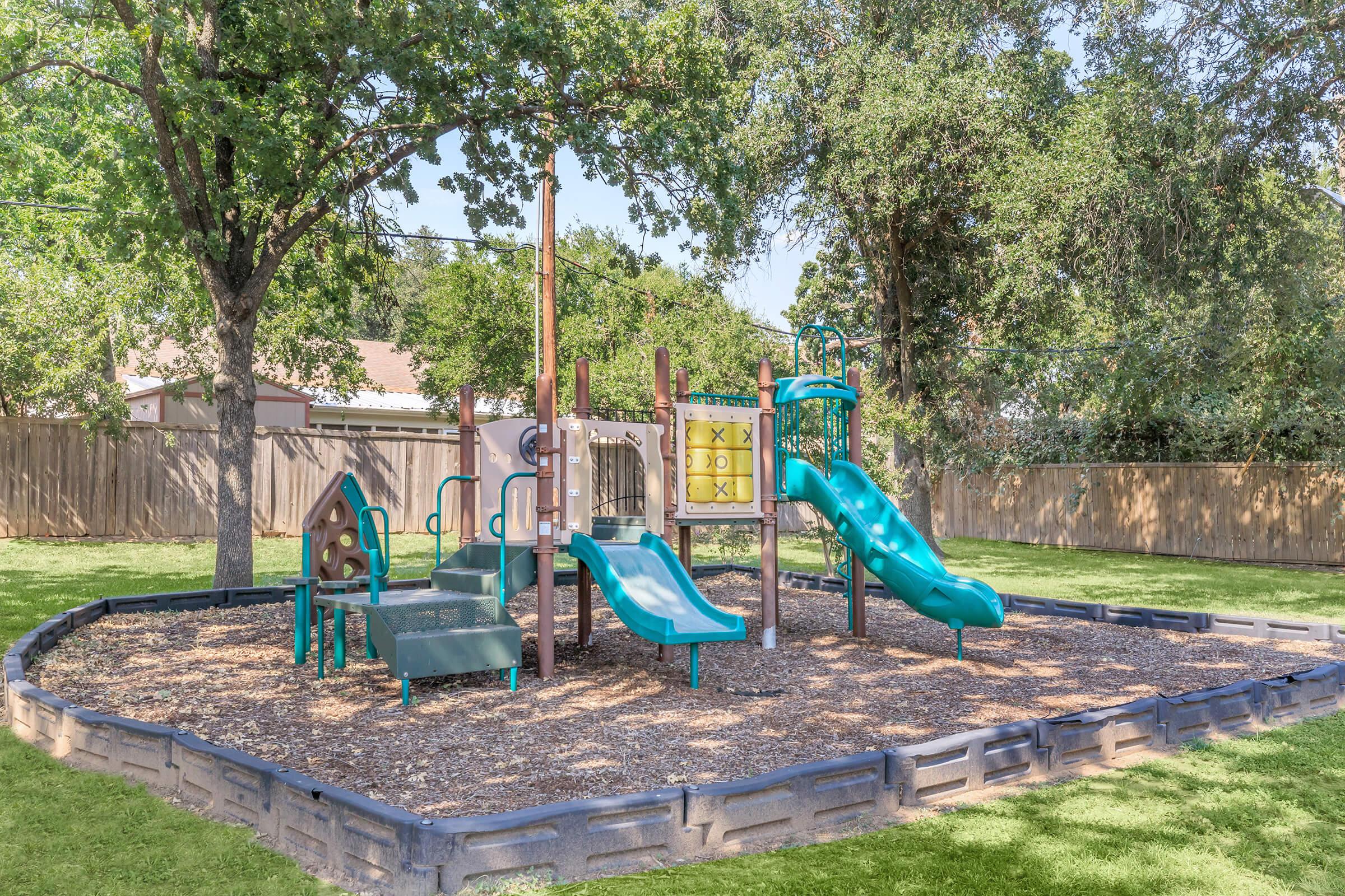A playground featuring two slides, climbing structures, and swings, surrounded by grass and trees. The play area is bordered by a low wall and contains mulch on the ground for safety. Ideal for children to play and socialize.