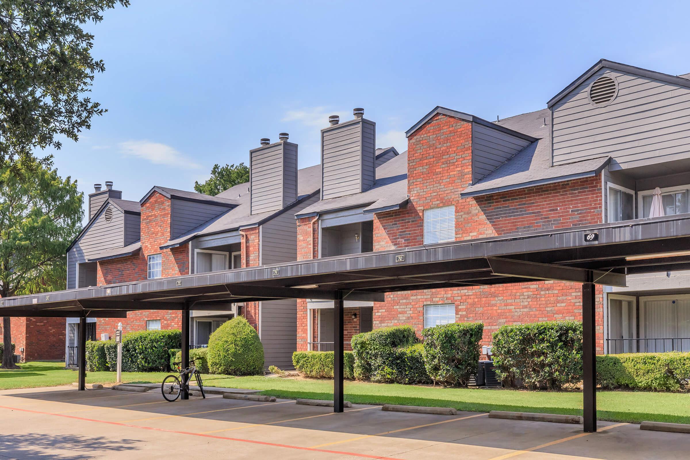 A view of a multi-level apartment complex featuring brick exteriors, sloped roofs, and multiple chimneys. The building has covered parking spaces in the foreground, with neatly trimmed shrubs and grassy areas. Blue sky and trees are visible in the background.