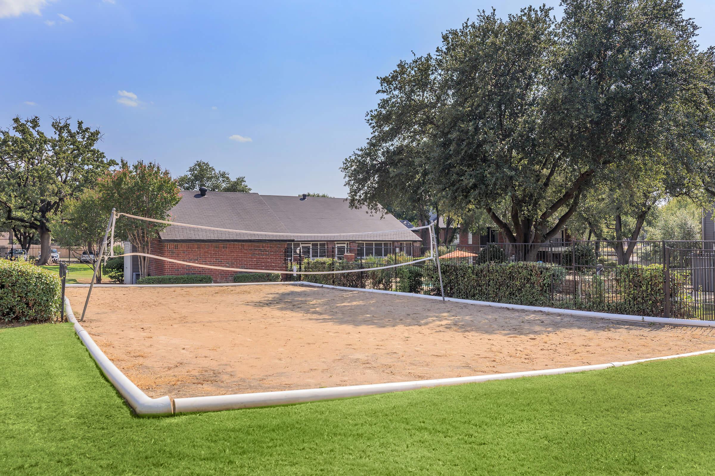 A sandy volleyball court surrounded by greenery, with a house visible in the background. The court features a net and is enclosed by a fence, under a clear blue sky. The area is well-maintained with grass in the foreground and trees providing shade.