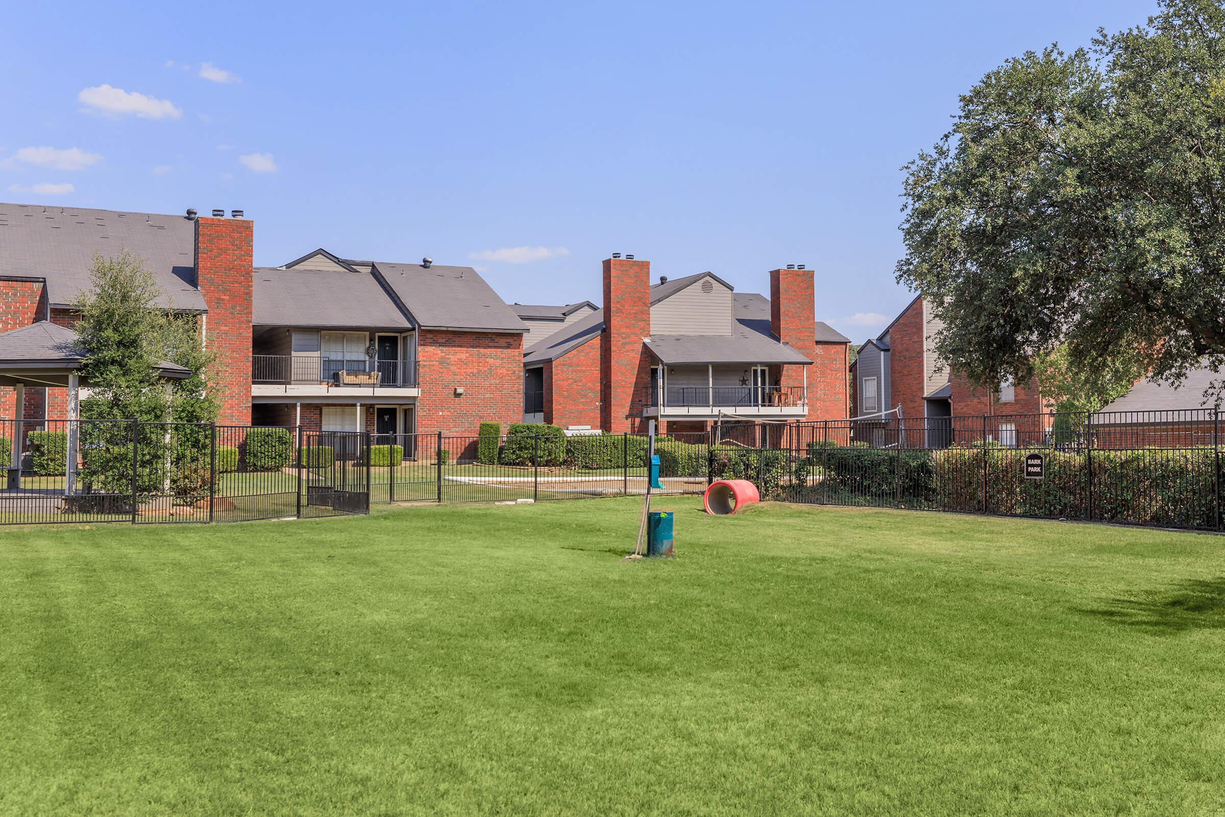 A well-maintained grassy area with a fence, surrounded by brick apartment buildings. In the foreground, there is a playground structure and a large red tube. The sky is clear with a few clouds, and trees are visible in the background, enhancing the outdoor space.
