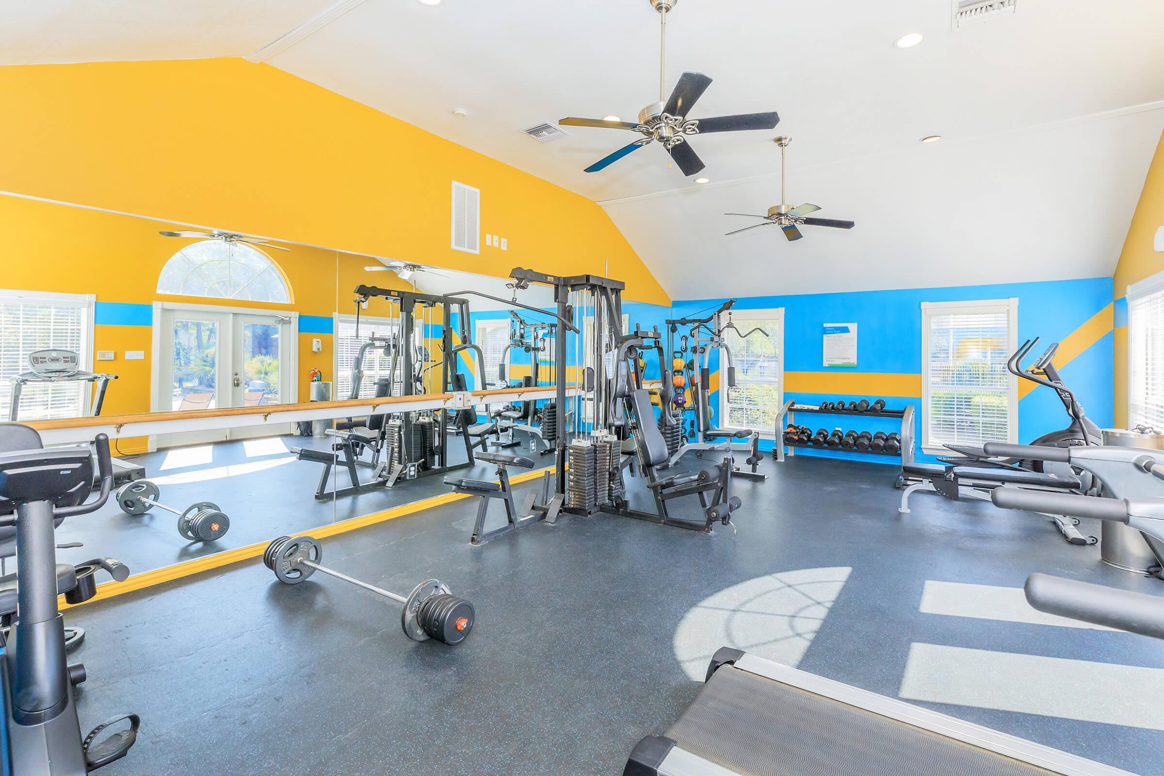 A bright and spacious gym featuring various workout equipment, including a weight machine, dumbbells, a treadmill, and a stationary bike. The walls are painted in blue and yellow, and large windows let in natural light, creating an inviting atmosphere for exercise.