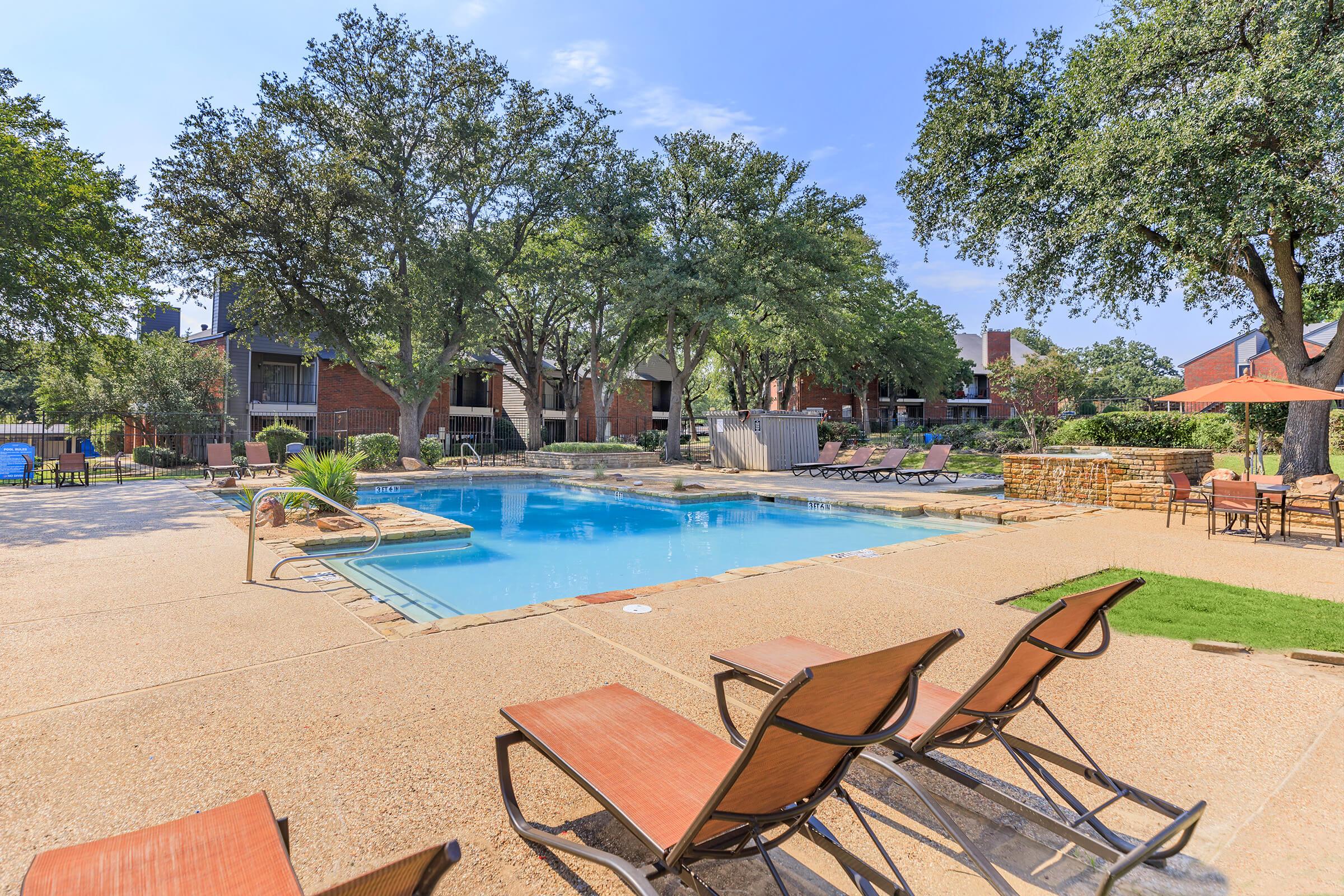 A spacious outdoor pool area surrounded by lounge chairs and trees. The pool is clear and inviting, with a stone border. In the background, there are multi-story residential buildings and additional seating areas, creating a relaxing atmosphere. The sky is blue and sunny.
