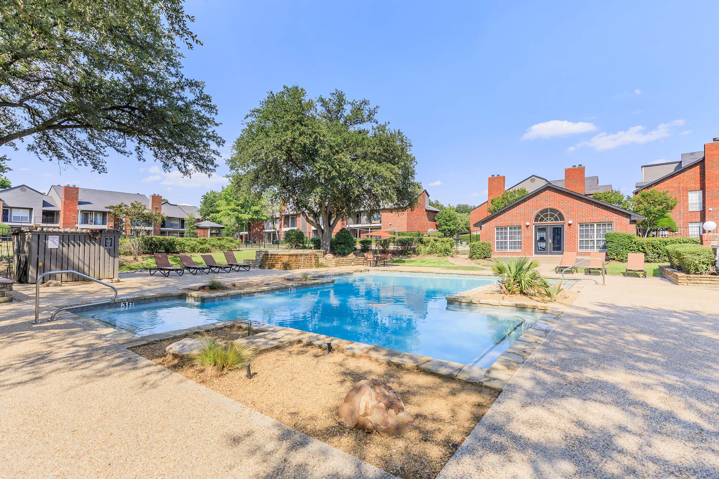 A tranquil pool area surrounded by lush greenery, featuring lounge chairs and a landscaped garden. The pool is clear and inviting, with a stone border. Nearby, two red-brick buildings are visible, and the sky is bright blue with a few clouds, contributing to a relaxing outdoor atmosphere.