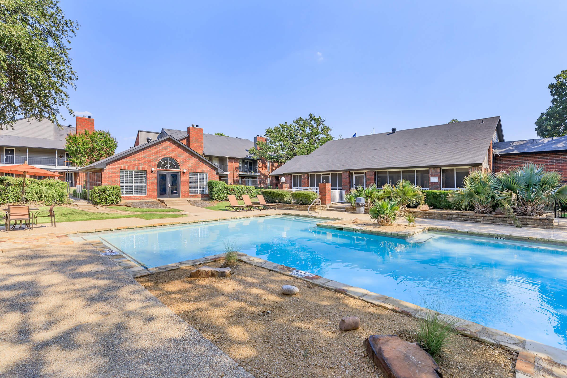 A serene outdoor pool area surrounded by lush greenery and landscaped gardens. Lounge chairs are positioned by the poolside, and a few umbrellas provide shade. Nearby, there are adjacent buildings with a brick façade, creating a welcoming atmosphere for relaxation and leisure.