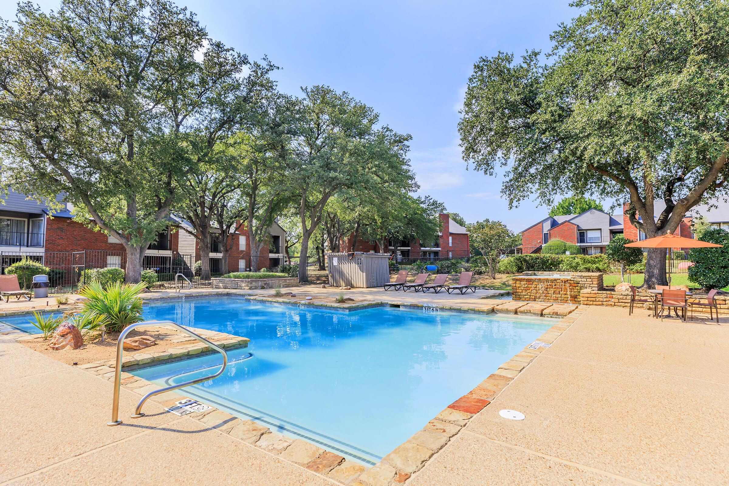 A serene outdoor swimming pool surrounded by lush green trees and lounge chairs. The pool area features a stone edge, with patio tables and umbrellas for shade, creating a relaxing atmosphere in a residential setting. Bright blue skies enhance the inviting scene.