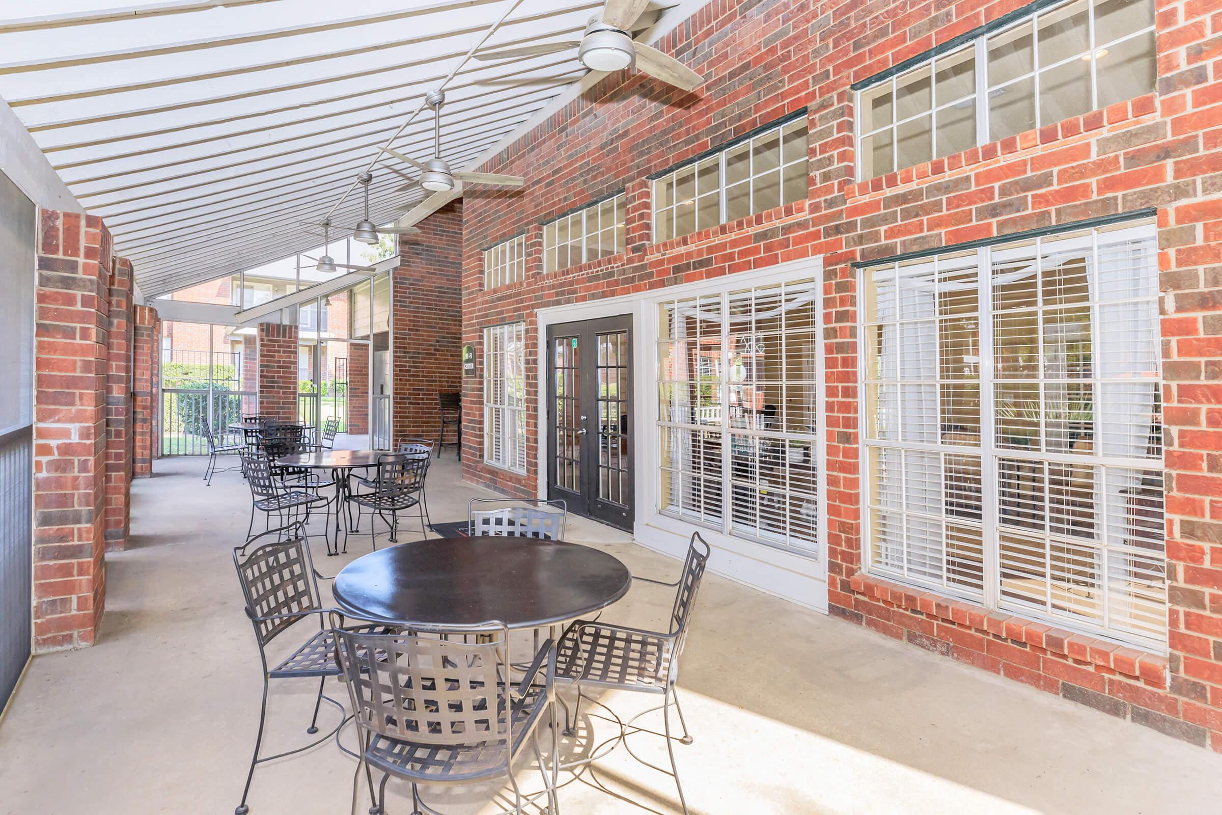 An outdoor seating area featuring black metal tables and chairs under a covered patio with brick walls and large windows. The space is bright and airy, designed for relaxation or gatherings.