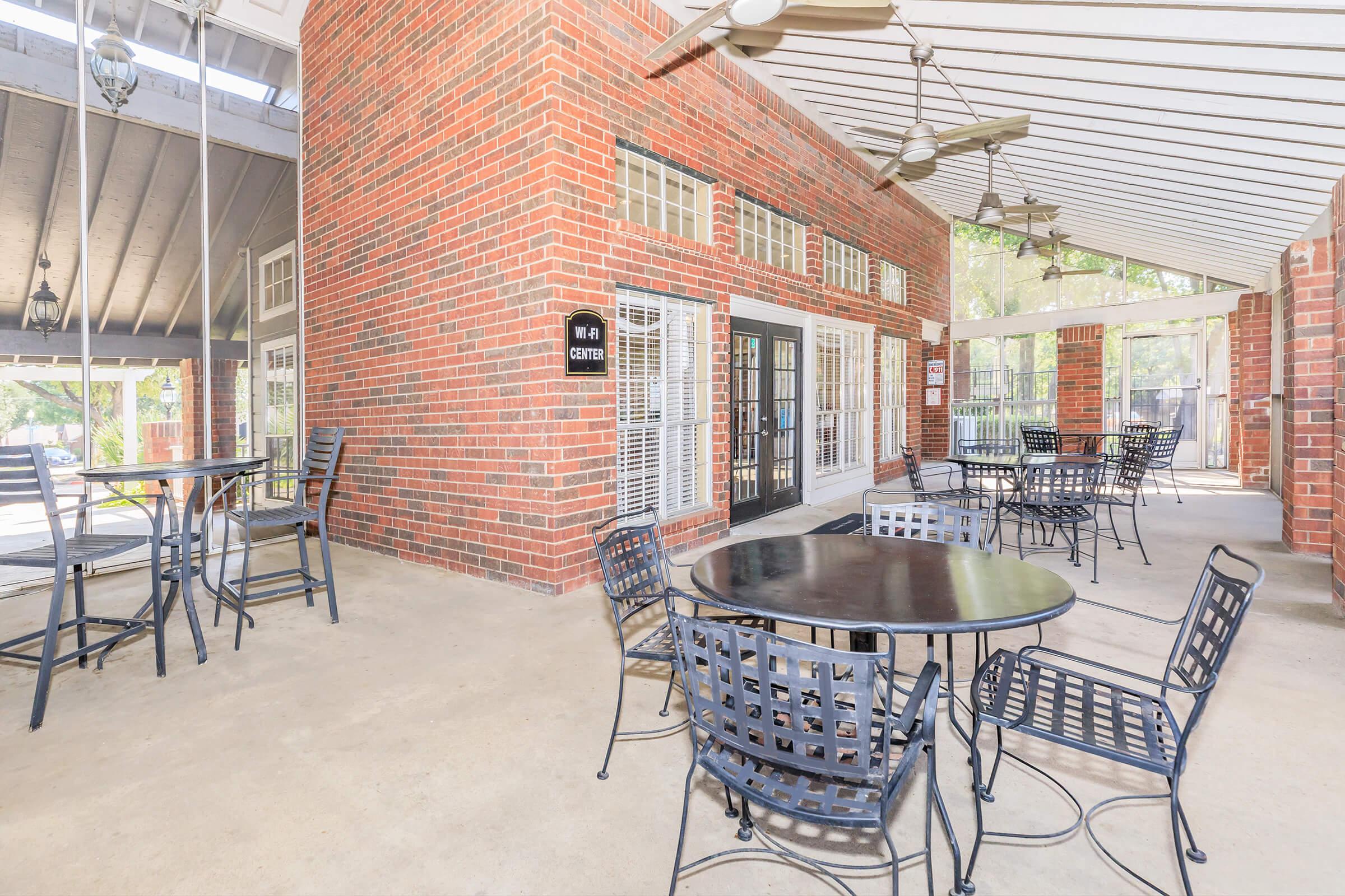 A spacious outdoor seating area featuring black metal tables and chairs, surrounded by red brick walls and large windows. The space is covered by a roof, and there are ceiling fans for ventilation, creating a comfortable atmosphere suitable for gatherings or relaxation.