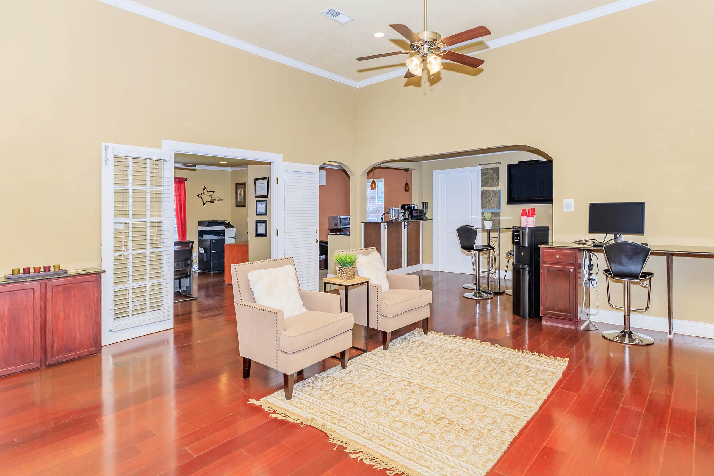 A spacious living room featuring two beige armchairs on a patterned rug. The room has wooden flooring, beige walls, and a ceiling fan. To the left, there's an open archway leading to a kitchen area with dark cabinetry and modern appliances. A small workspace with a computer is visible in the corner.