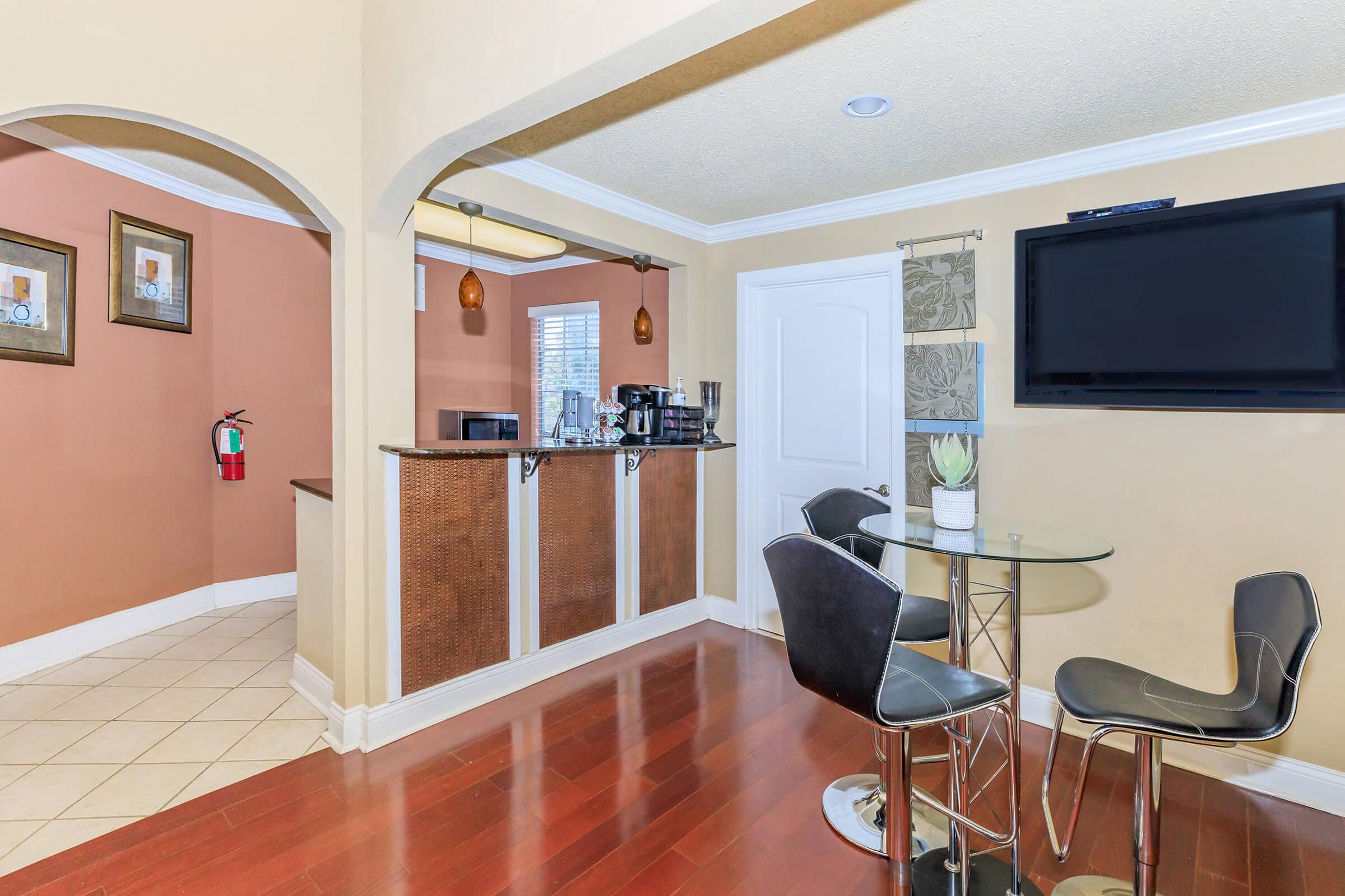 Modern kitchen space featuring a bar area with a glass table and black stools. The walls are painted in warm tones, with framed artwork and a fire extinguisher visible. A microwave and coffee maker are on the counter, and a flat-screen TV is mounted nearby. The flooring is wooden, creating a cozy atmosphere.