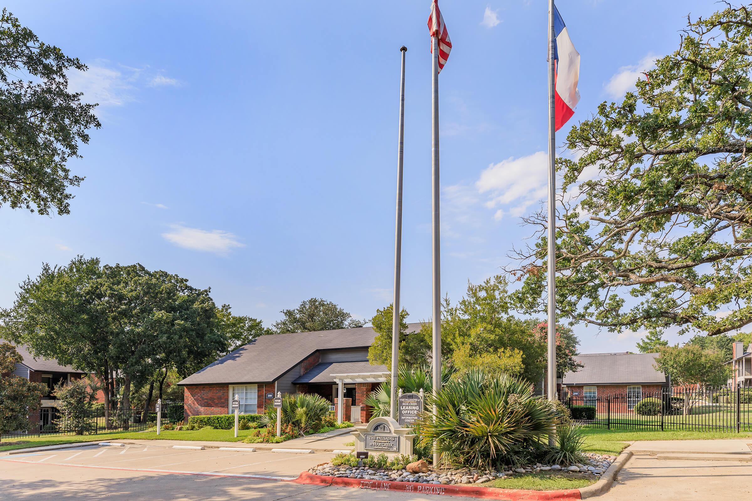 A brick building with a sloped roof, surrounded by trees and landscaping. In front, two flags are raised on flagpoles. A sign is displayed in the foreground, and the area appears well-maintained with a clear blue sky overhead.