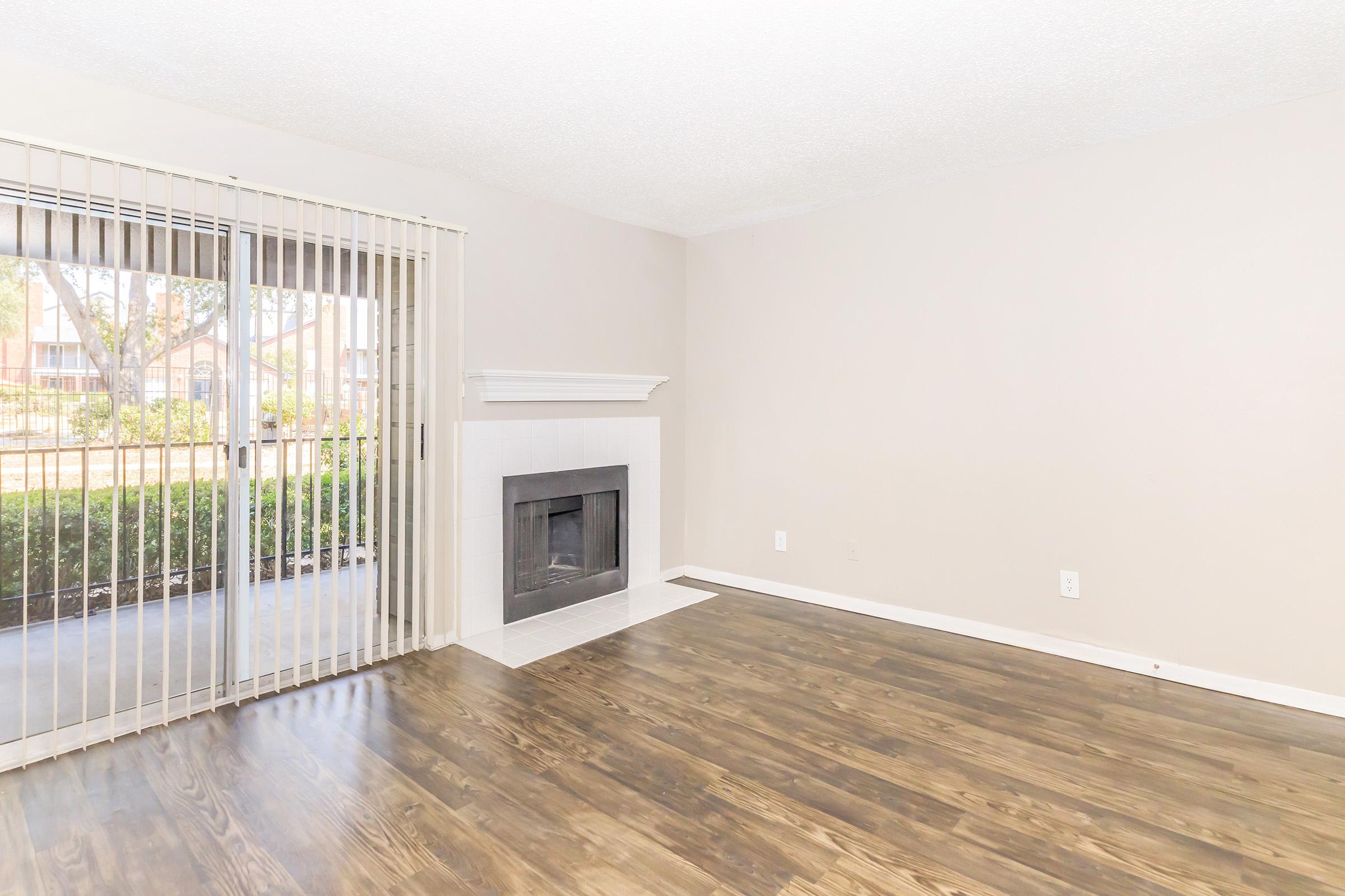 A light and airy living room featuring a large window with vertical blinds, a white fireplace, and hardwood-style flooring. The walls are painted in a soft beige color, creating a neutral and inviting atmosphere. The space is unfurnished, emphasizing its open layout.
