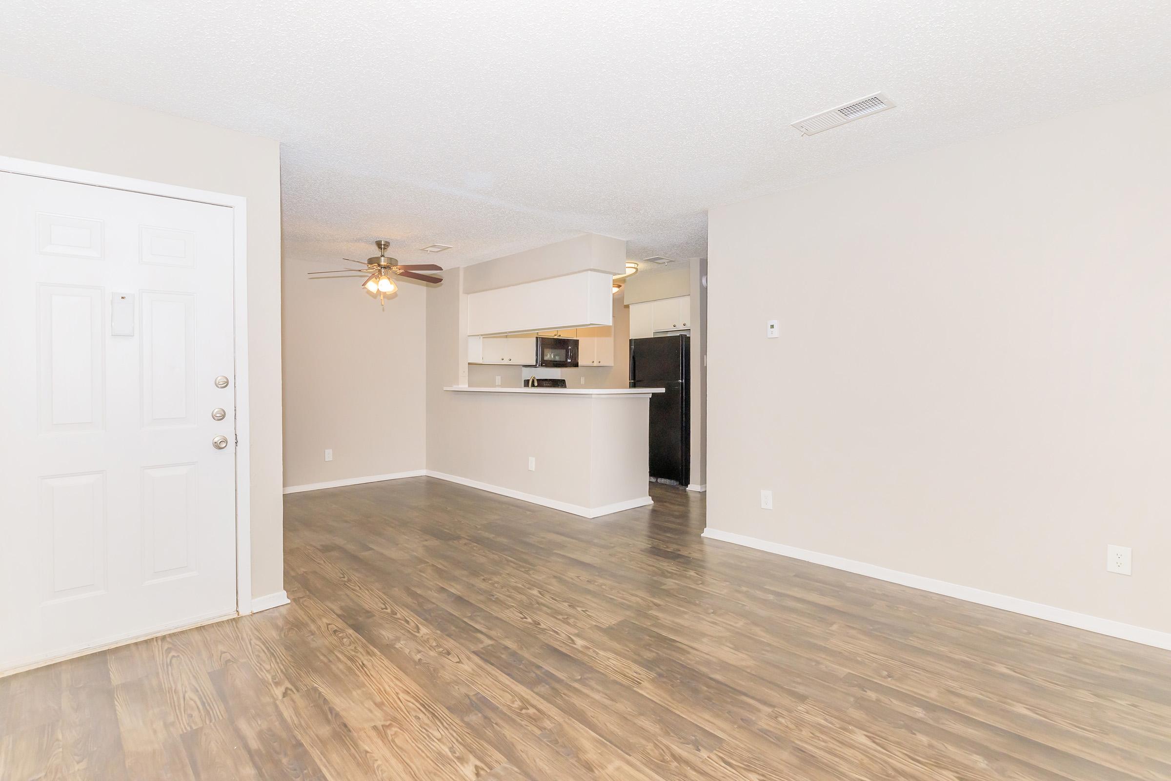 A spacious, unfurnished living room featuring light-colored walls, laminate flooring, and a ceiling fan. There's an entrance on the left and an open kitchen area visible in the background, complete with a breakfast bar. The overall ambiance is bright and inviting, suitable for personal decoration.