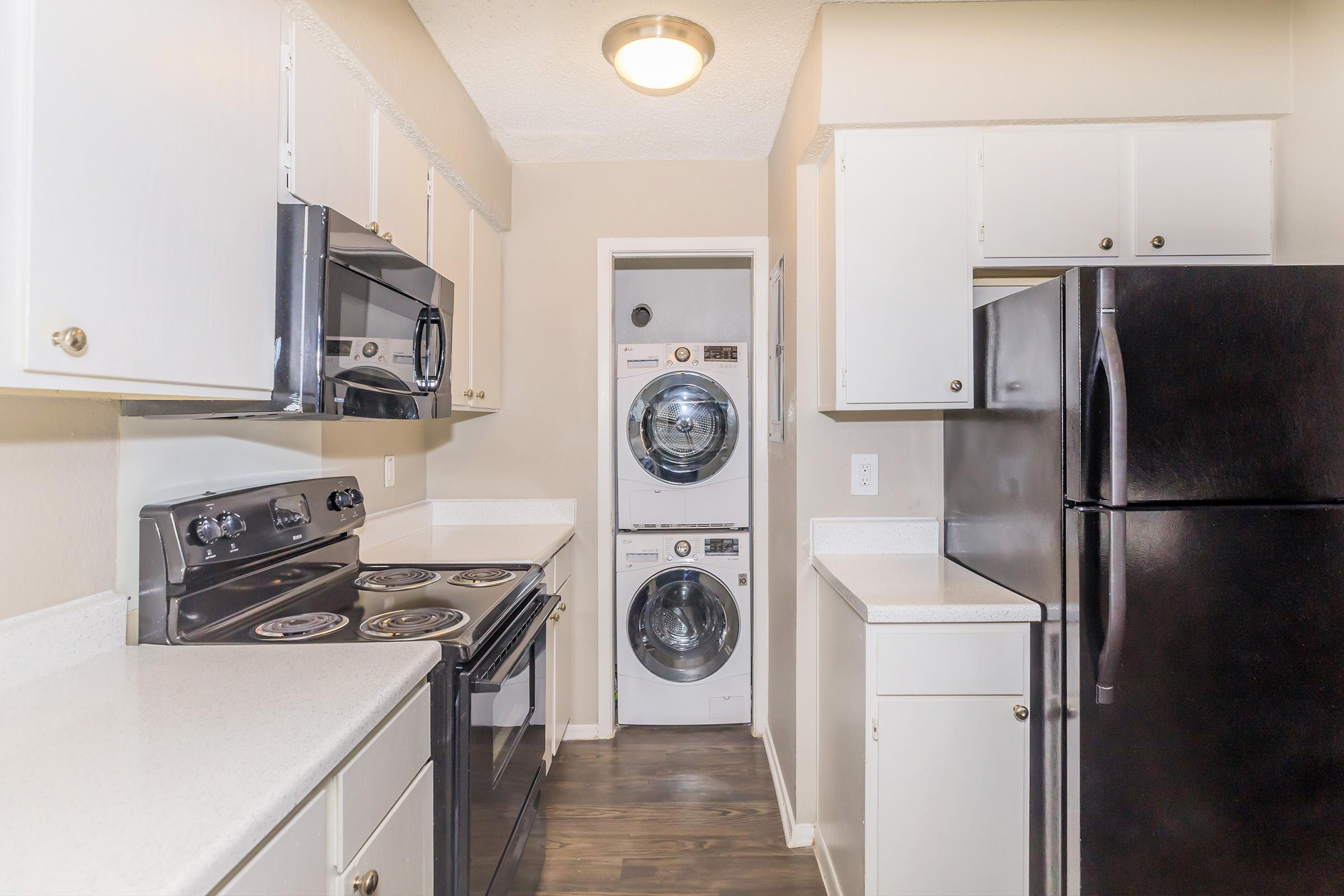 A modern kitchen featuring white cabinetry, stainless steel appliances, and a black refrigerator. The room has a stovetop and microwave, with a laundry area visible in the background, showcasing stacked washer and dryer units. The overall design is clean and functional, with a light color scheme.