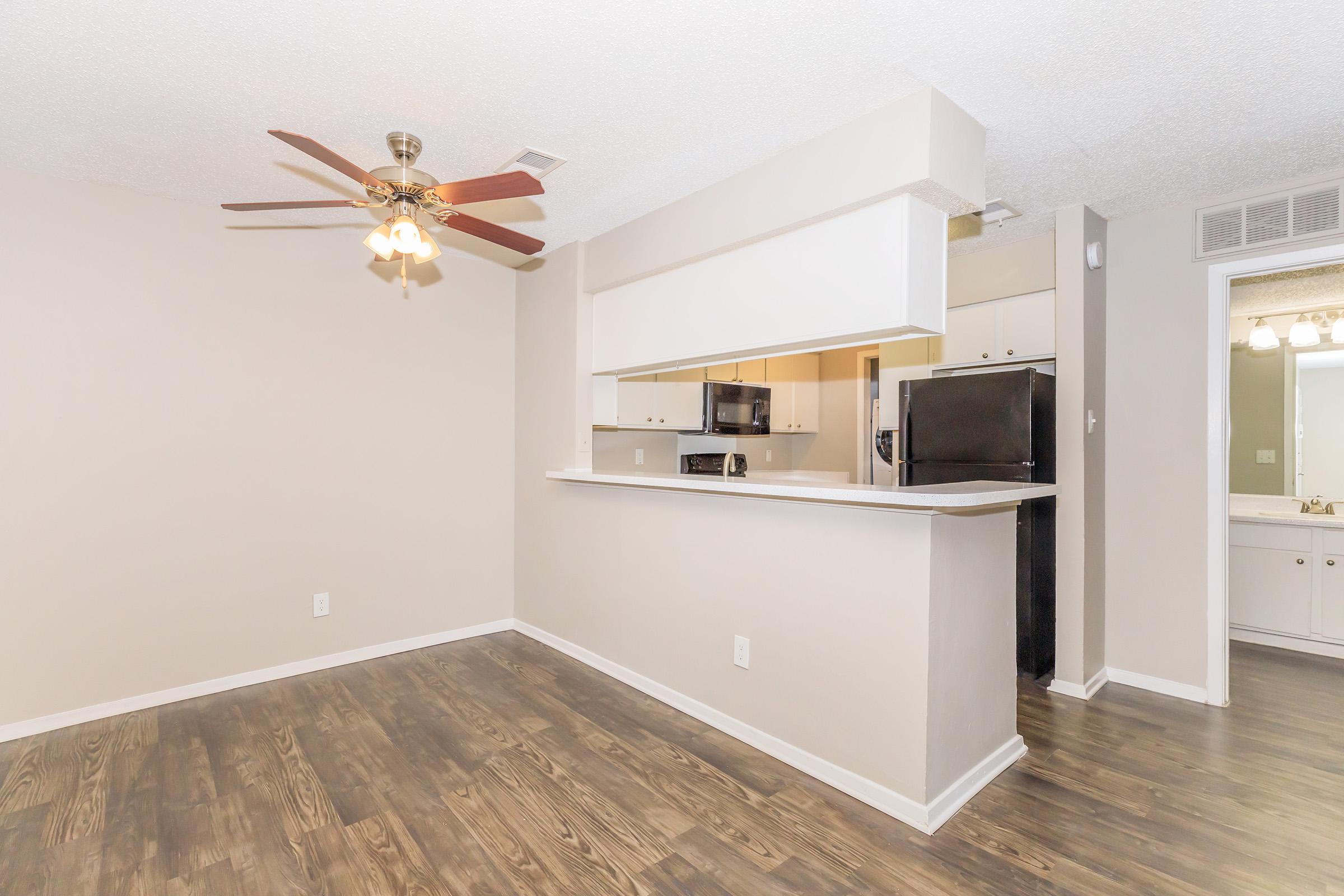 A modern living area featuring a ceiling fan, light-colored walls, and hardwood-style flooring. There is a breakfast bar with a view into a kitchen equipped with black appliances. The layout is open and spacious, creating a bright and inviting atmosphere.