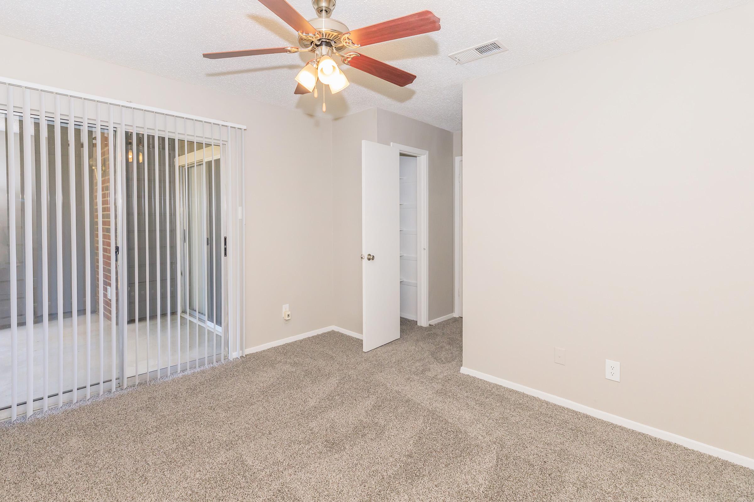 A spacious room with beige walls and plush carpet. A ceiling fan with wooden blades hangs from the ceiling. Light streams through sliding glass doors with vertical blinds leading to a patio. A doorway opens to a small closet, enhancing the room's functionality. The overall atmosphere is bright and inviting.