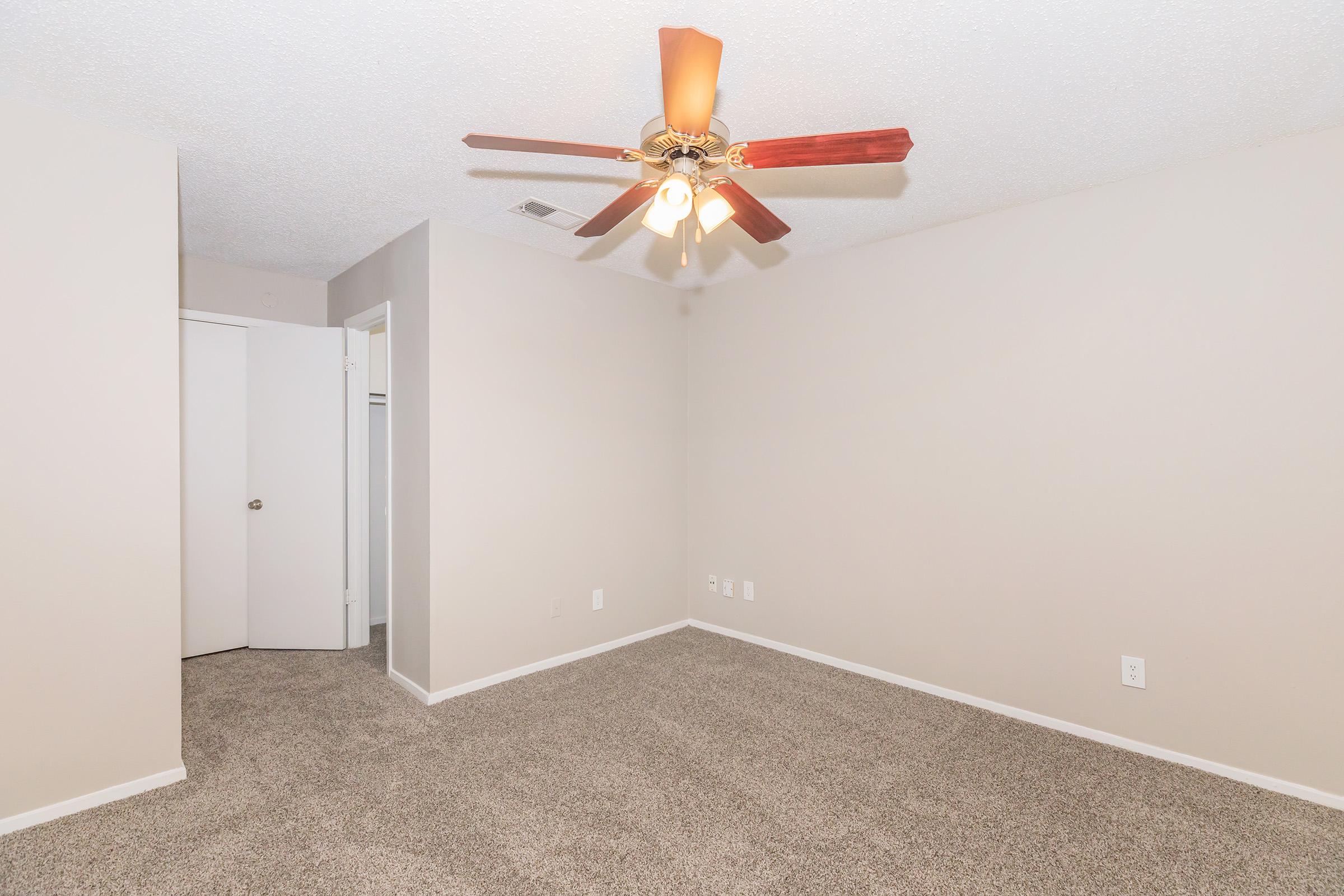 A spacious room featuring light beige walls and plush carpet. A ceiling fan with wooden blades is mounted in the center, providing ventilation. To one side, there is a closet with a white door, and another door leads outside of the room. The overall atmosphere is bright and inviting, perfect for various uses.