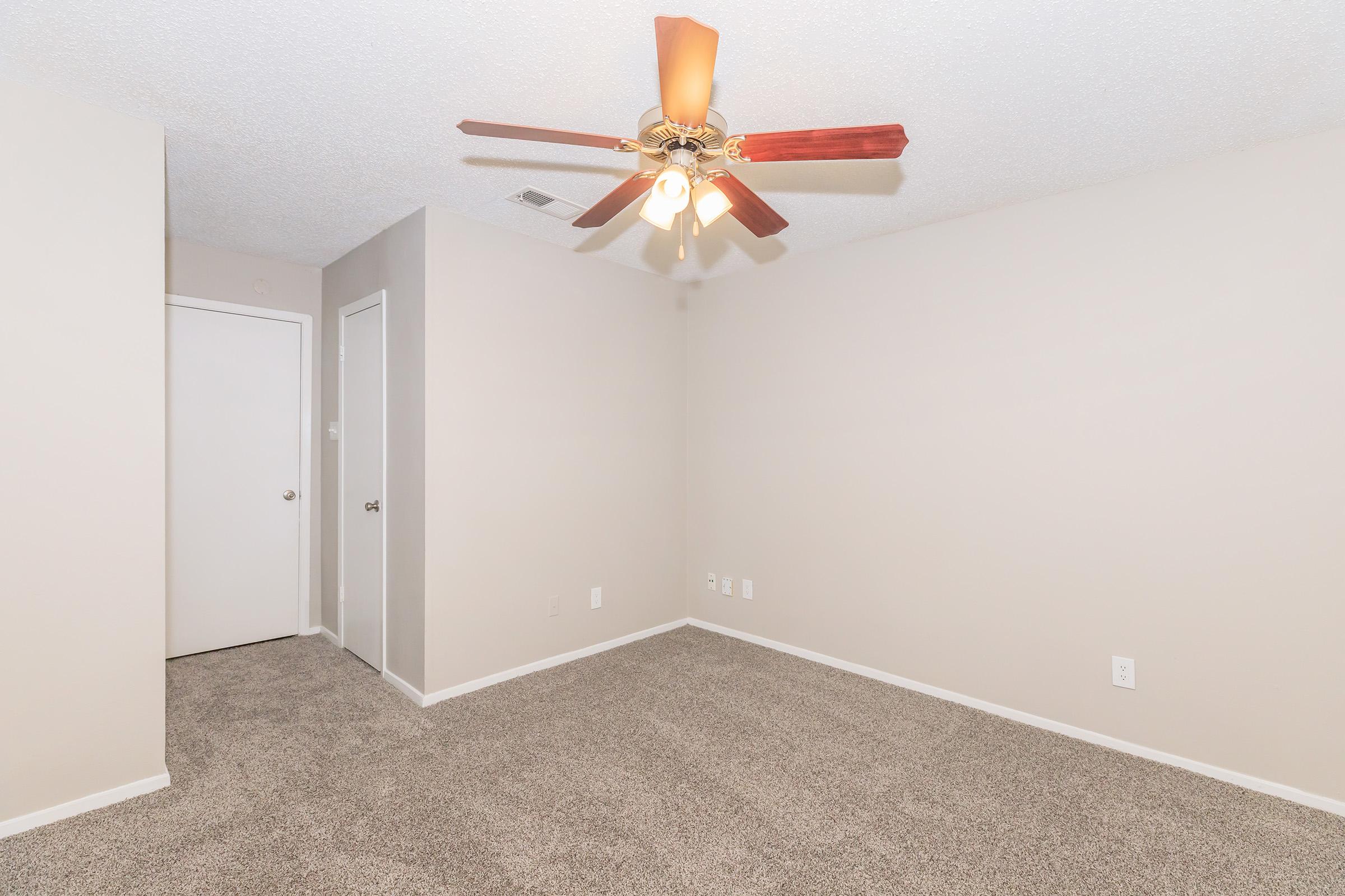 A spacious, empty room with light beige walls and a ceiling fan with wooden blade accents. The floor is covered in light brown carpet. There is a closed door on the left and a closet door on the right, creating a clean and minimalist look, ideal for personalization.