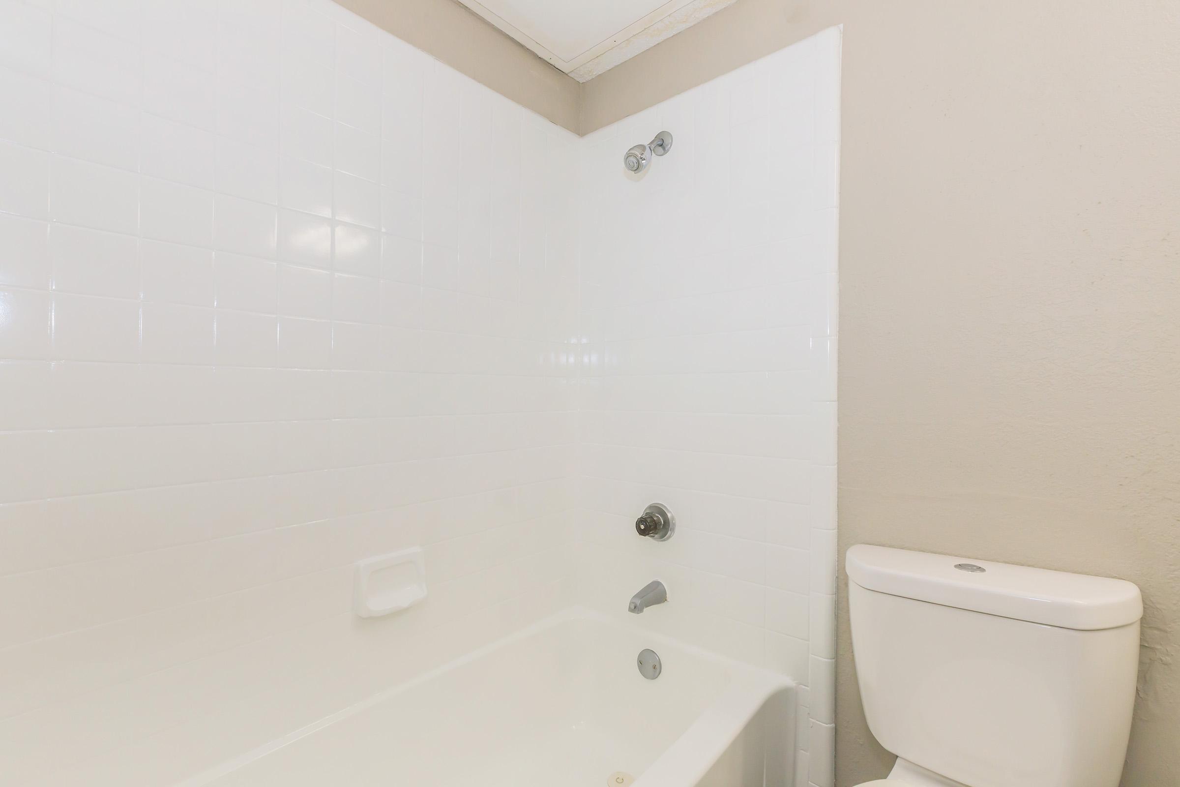 A clean and minimalist bathroom featuring a white tiled wall, a bathtub with a chrome faucet and showerhead, and a white toilet. The walls are a light beige, providing a neutral backdrop.