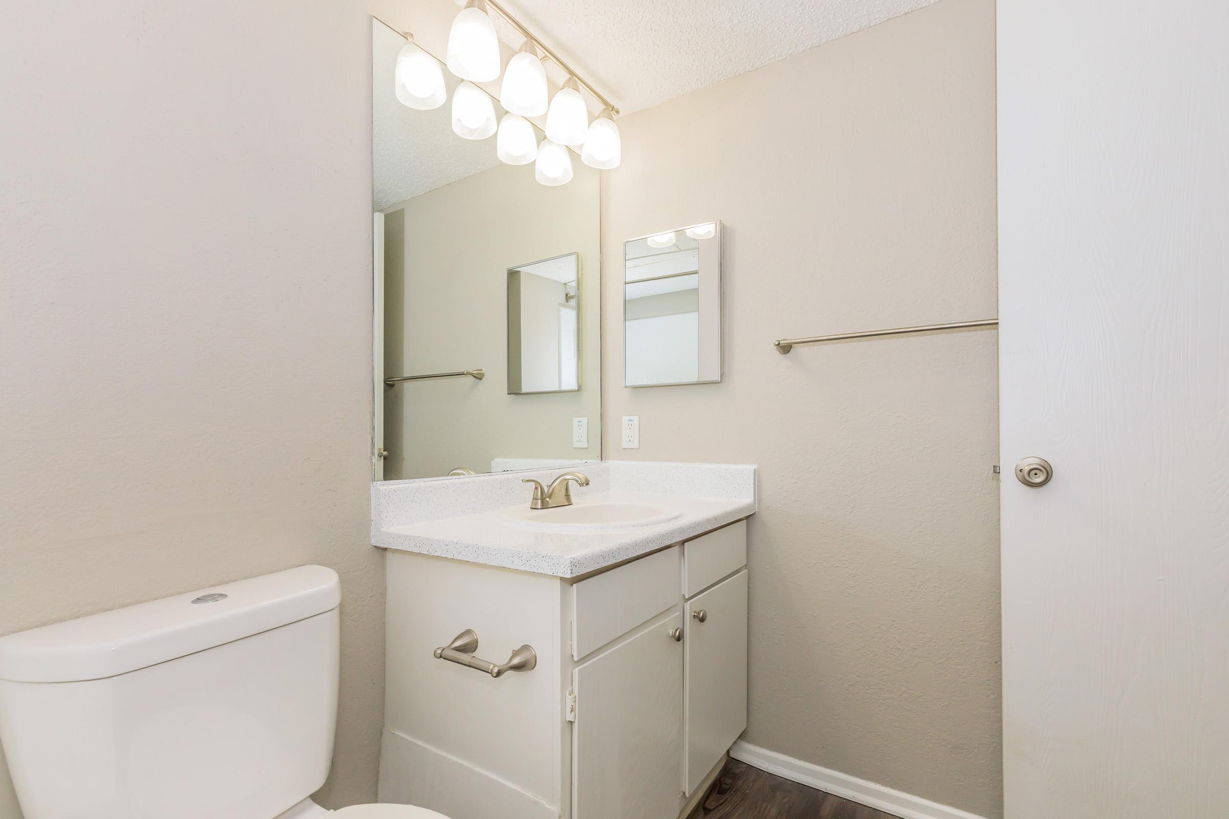 A modern bathroom featuring a white toilet and a light-colored countertop with a sink. Two mirrors are mounted above the countertop, and there are light fixtures above them. The walls are painted in a neutral tone, and a towel bar is visible beside the countertop. A door leads out of the space.