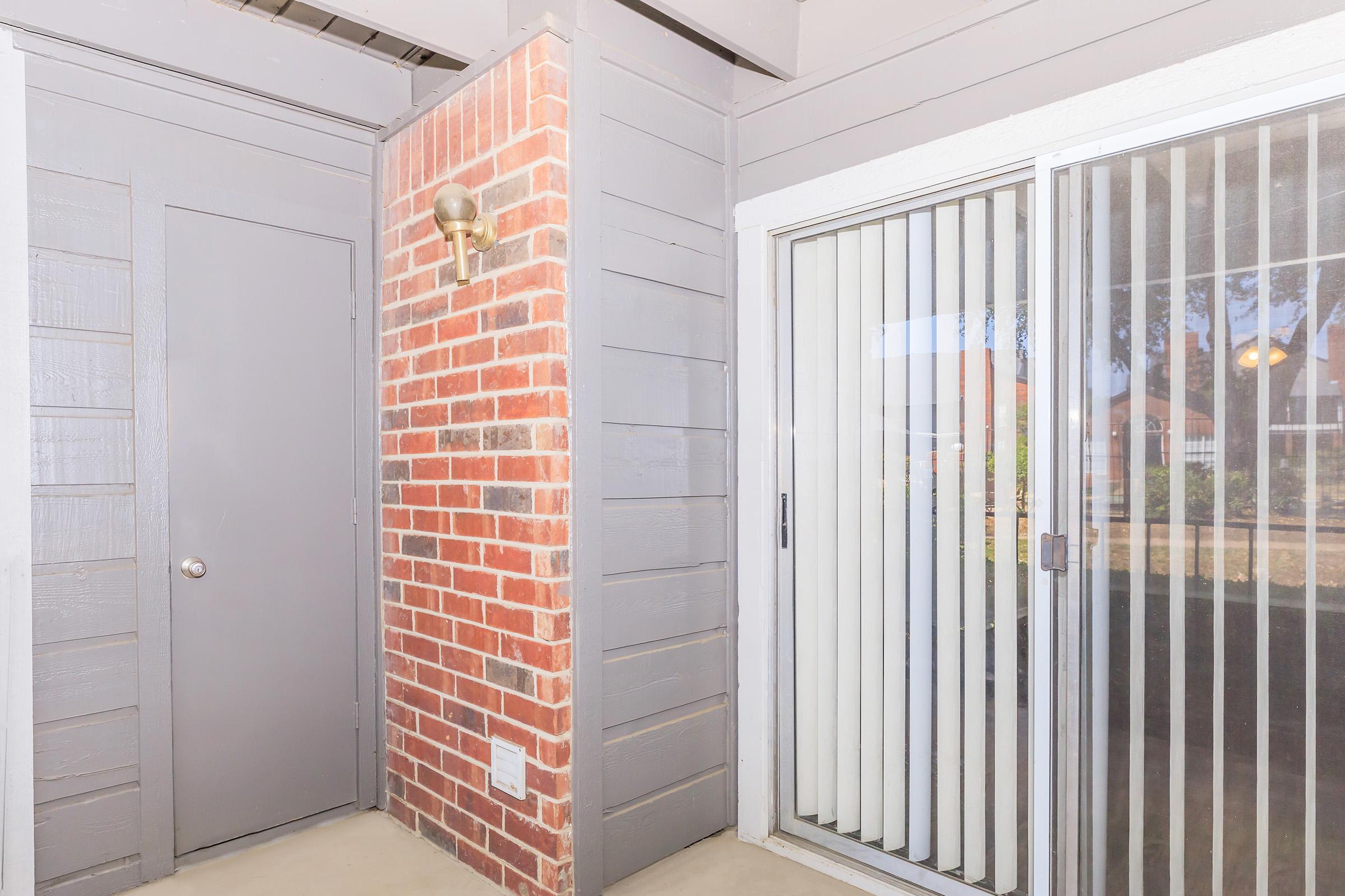 Interior corner of an apartment with a brick wall and gray wooden paneling. A closed door is on one side, and a sliding glass door with vertical blinds is on the other. The area is well-lit with a wall-mounted light fixture, creating a cozy entryway feel.