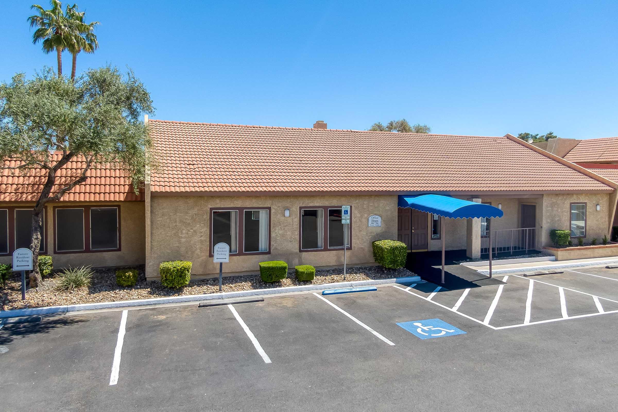 A single-story building with a tiled roof and several windows. There are parking spaces in front, including a designated handicap spot. A blue awning covers the entrance, and there are shrubs and small trees in the landscape. The sky is clear and sunny.
