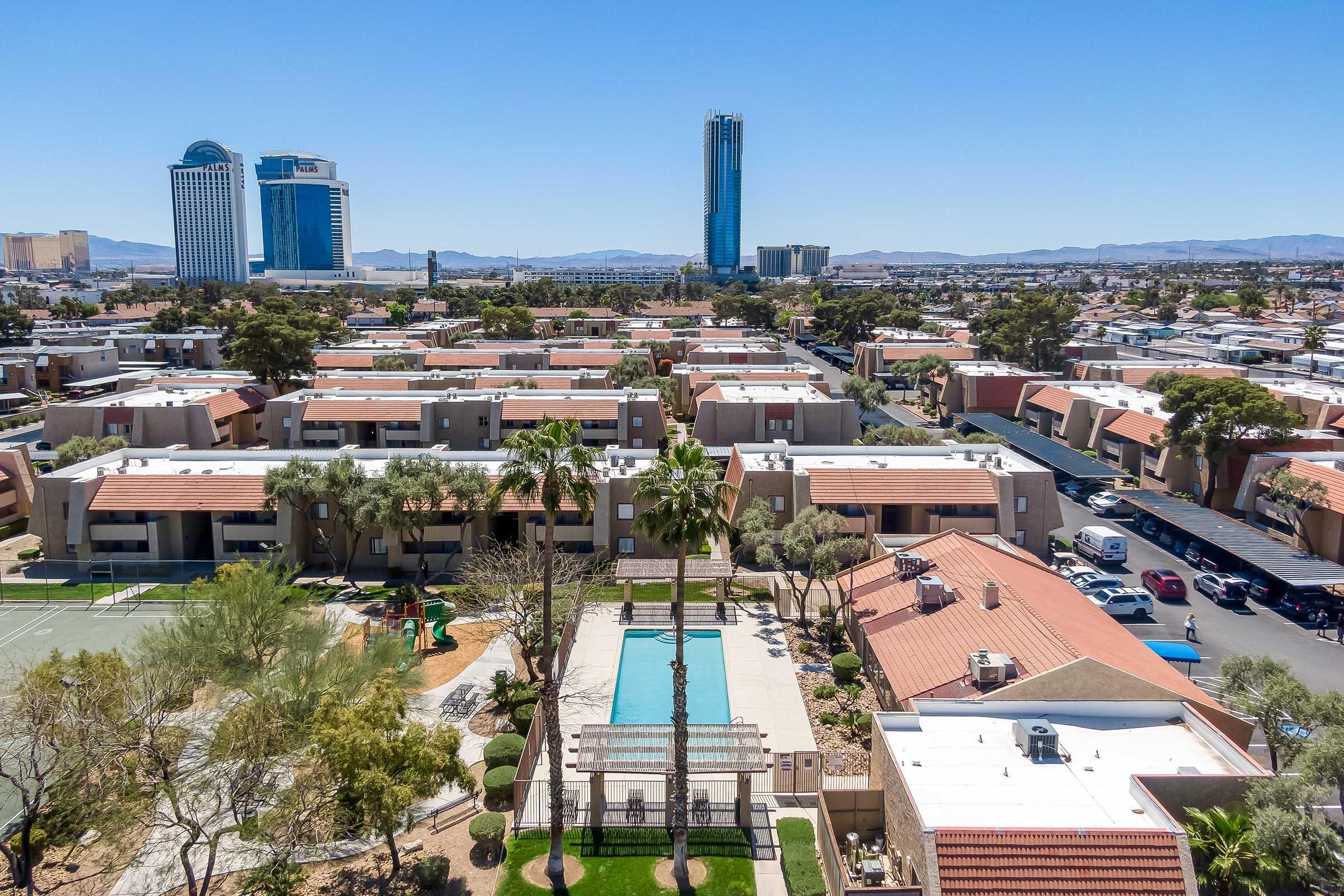 Aerial view of a residential area featuring multiple buildings with red roofs, a swimming pool surrounded by palm trees, and a nearby tennis court. In the background, tall modern skyscrapers and distant mountains are visible under a clear blue sky.