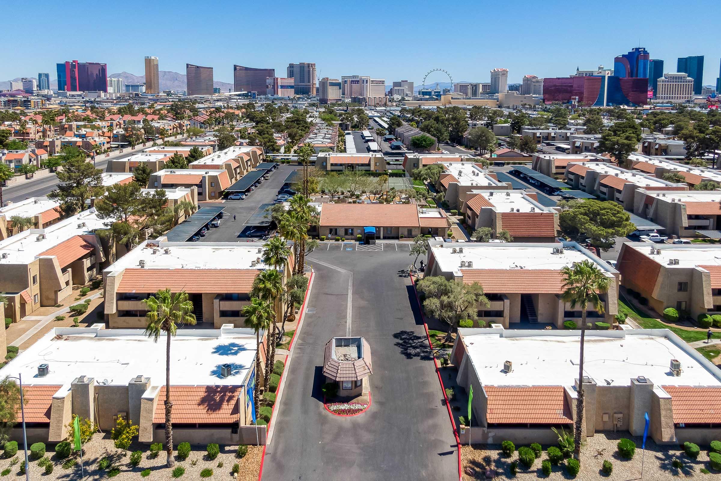 Aerial view of a residential area with single-story buildings and palm trees, leading to a street surrounded by parked cars. In the background, the Las Vegas skyline features various hotels and resorts, including notable structures and a Ferris wheel, under a clear blue sky.