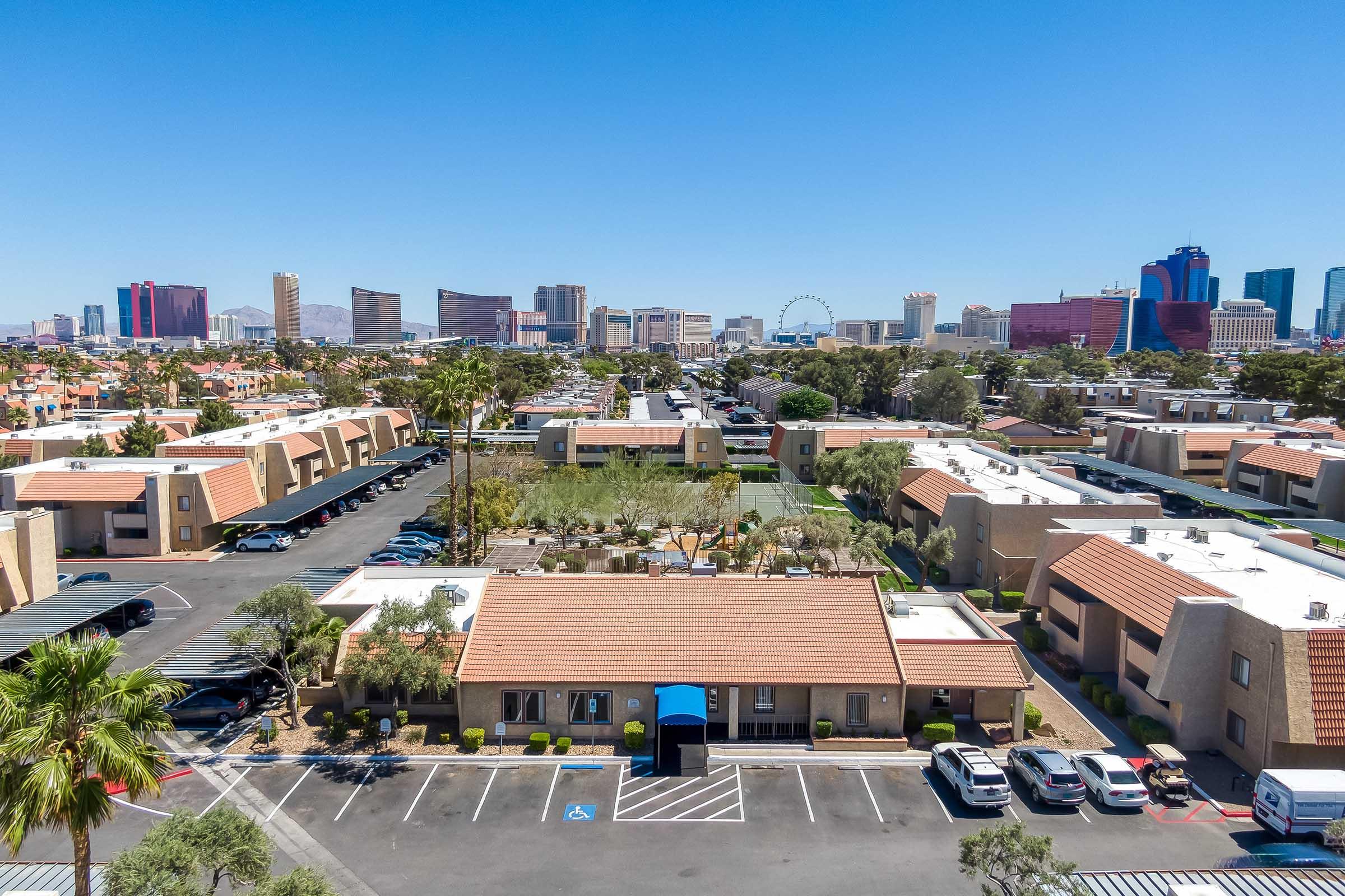 Aerial view of a suburban area with multi-story apartment buildings and a parking lot, framed by palm trees. In the distance, the skyline of a city featuring modern hotels and a Ferris wheel is visible under a clear blue sky.