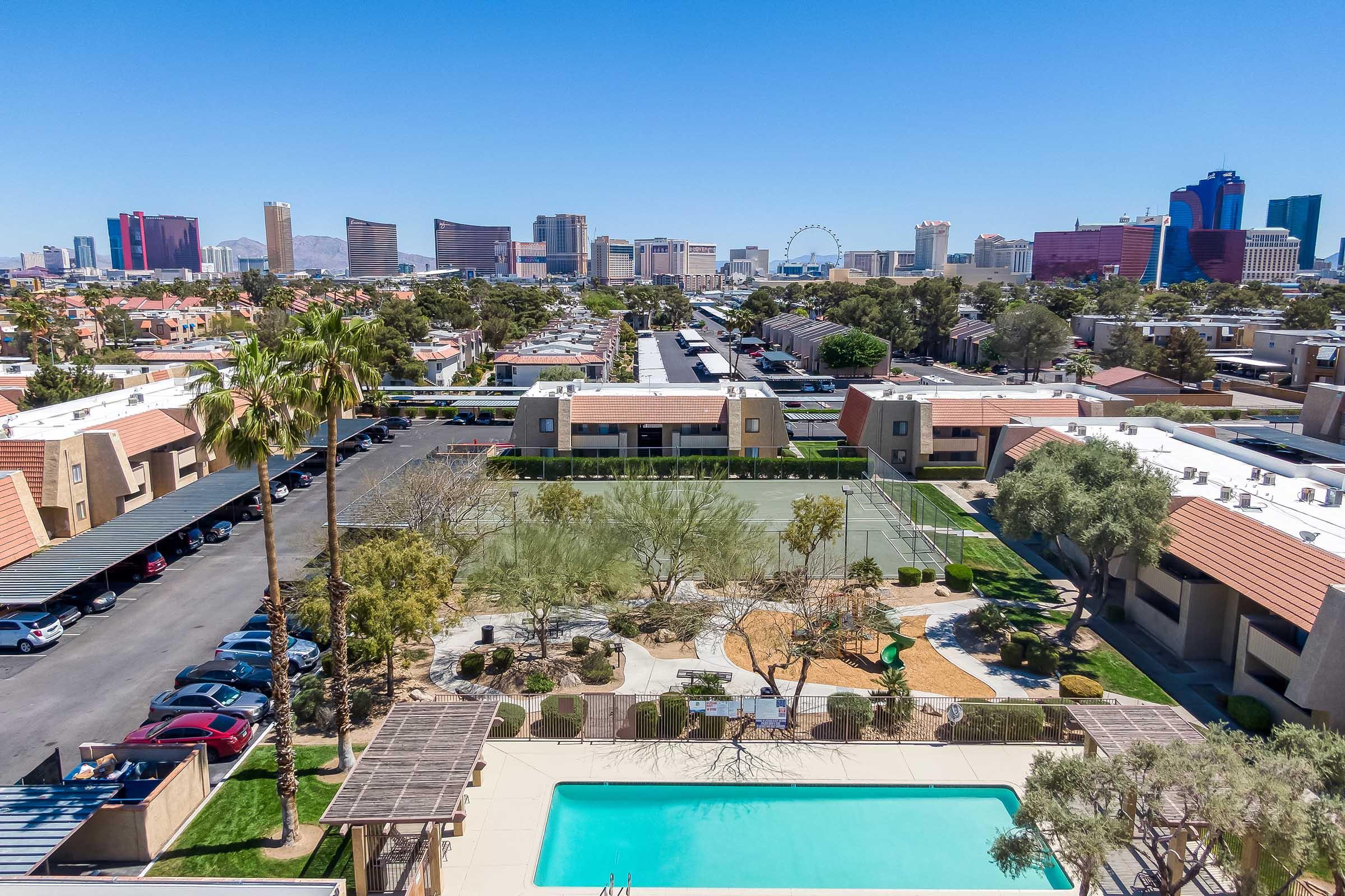 Aerial view of a residential area with multiple apartment buildings, a swimming pool, and landscaped gardens. In the background, the Las Vegas skyline features notable skyscrapers and a large Ferris wheel against a clear blue sky. Palm trees are visible in the foreground.