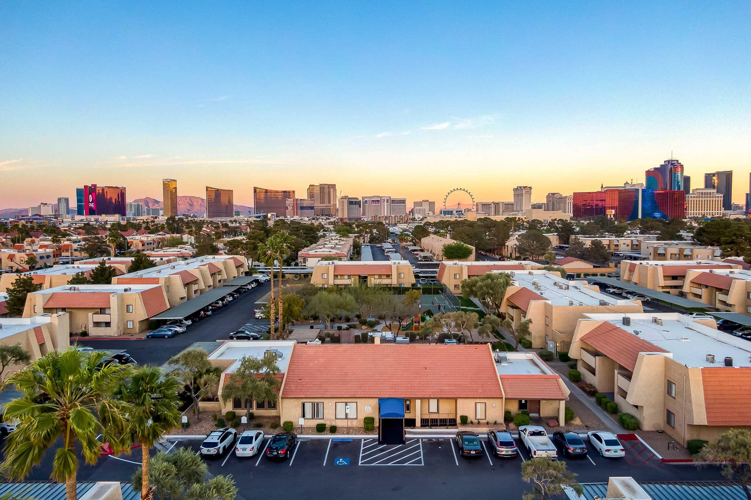 Aerial view of a residential area with low-rise buildings and a parking lot in the foreground, overlooking a city skyline at dusk. The skyline features high-rise buildings and a large ferris wheel, with vibrant colors in the sky as the sun sets. Palm trees are visible in the foreground.