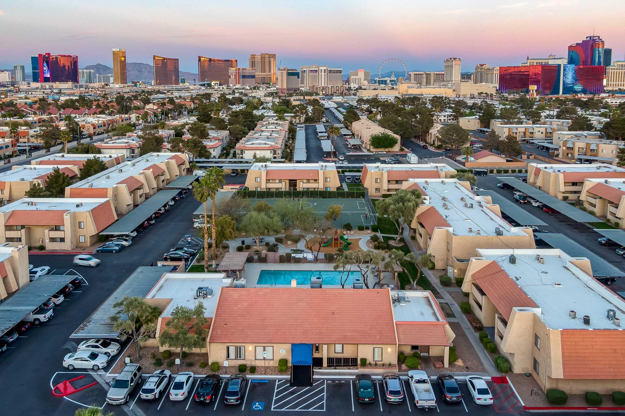 Aerial view of a residential area with apartment buildings, a swimming pool, and parked cars. In the background, the Las Vegas skyline features iconic hotels and casinos under a colorful sky at dusk. Palm trees are visible, along with a sports court, creating a vibrant suburban atmosphere.