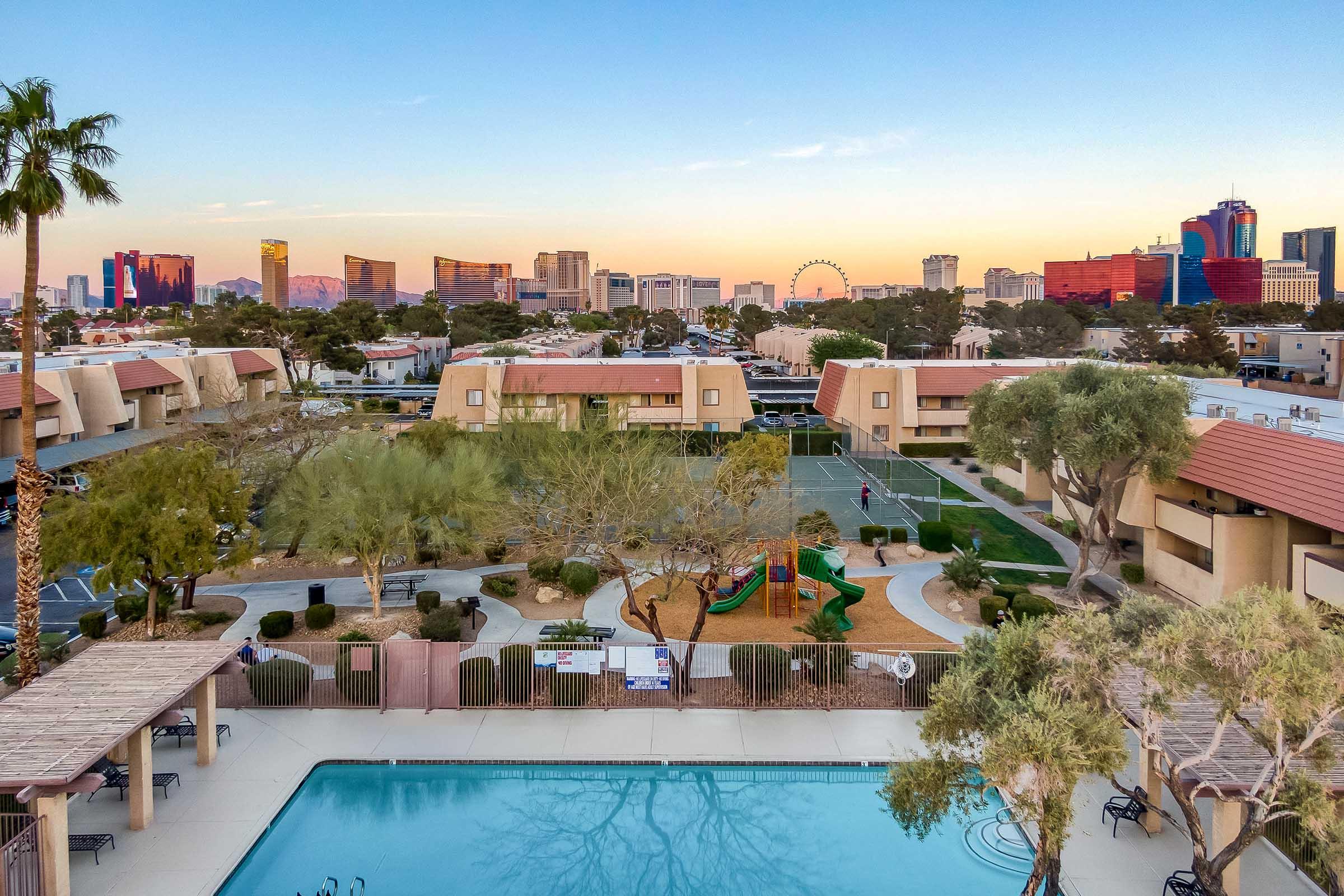 A view of a residential area featuring a swimming pool in the foreground, surrounded by landscaped gardens. In the background, the skyline of a city can be seen with tall buildings and a ferris wheel, under a colorful sunset sky.