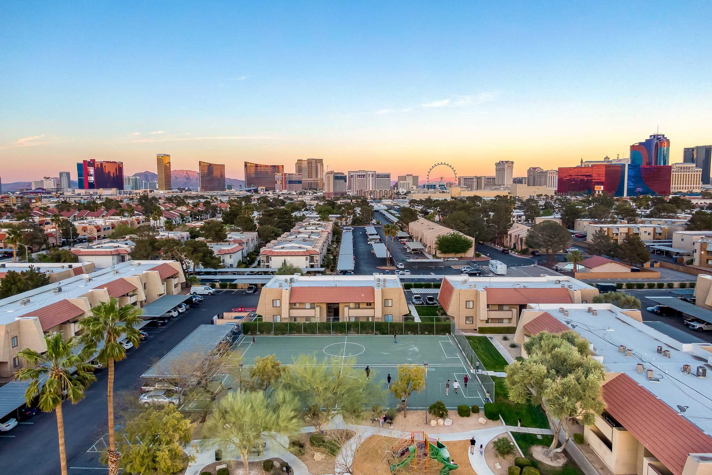 Aerial view of a suburban neighborhood with a basketball court and playground area in the foreground, contrasted against the Las Vegas skyline in the background. The cityscape features iconic buildings, illuminated by sunset hues, with palm trees and residential complexes visible below.