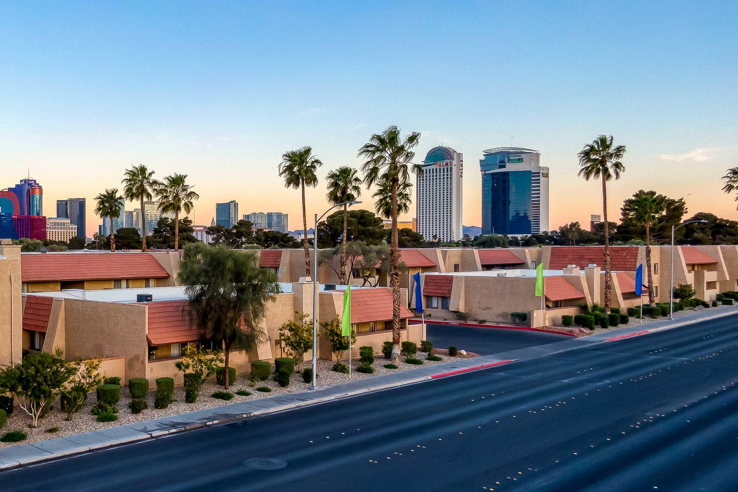 A cityscape view featuring palm trees lining a road, with low-rise buildings in the foreground and tall, modern skyscrapers in the background. The sky shows hints of sunset colors, casting a warm glow over the scene. The atmosphere is urban and inviting, typical of a sunny location.