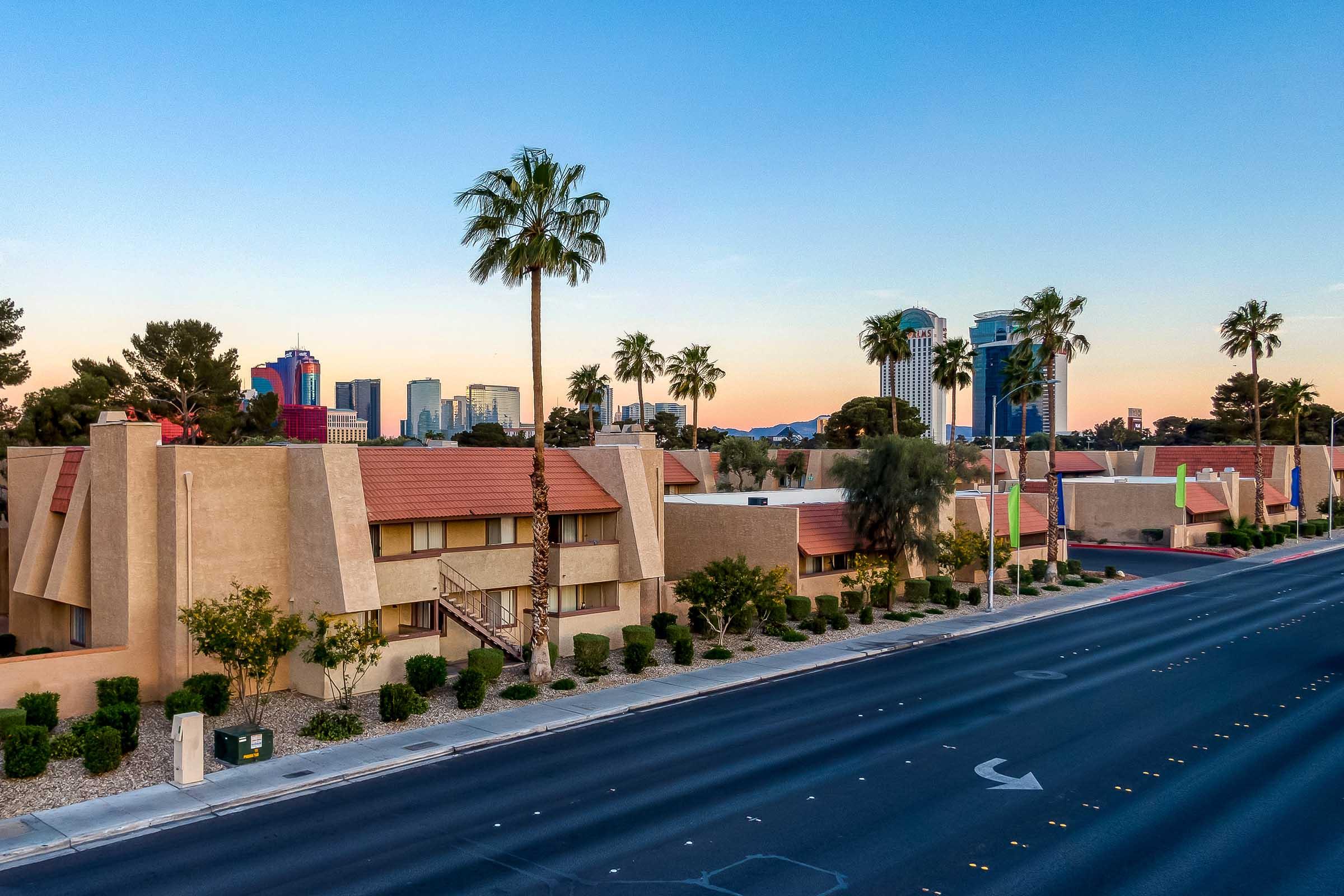 A view of a street lined with palm trees, leading towards a row of low-rise buildings with a desert landscape. In the background, the skyline of a city is visible, featuring modern skyscrapers against a colorful sunset sky.
