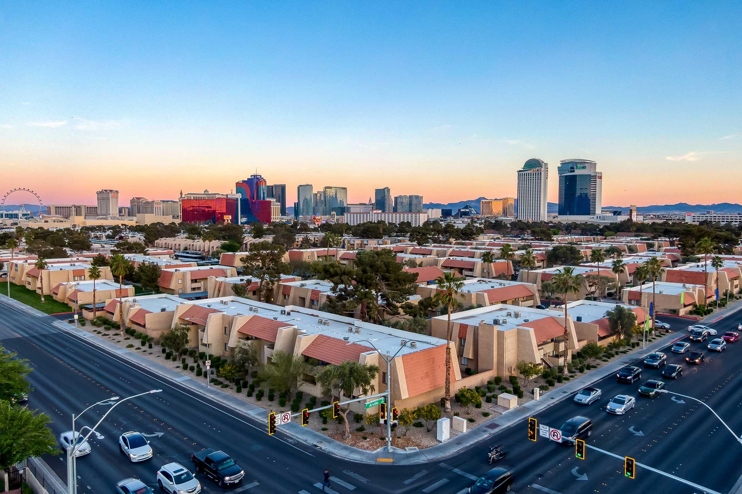 Aerial view of a Las Vegas neighborhood featuring low-rise buildings with red-tiled roofs, surrounded by palm trees. The backdrop showcases the Las Vegas Strip, highlighting various hotels and casinos against a colorful sunset sky. Busy streets with traffic and pedestrians are visible in the foreground.