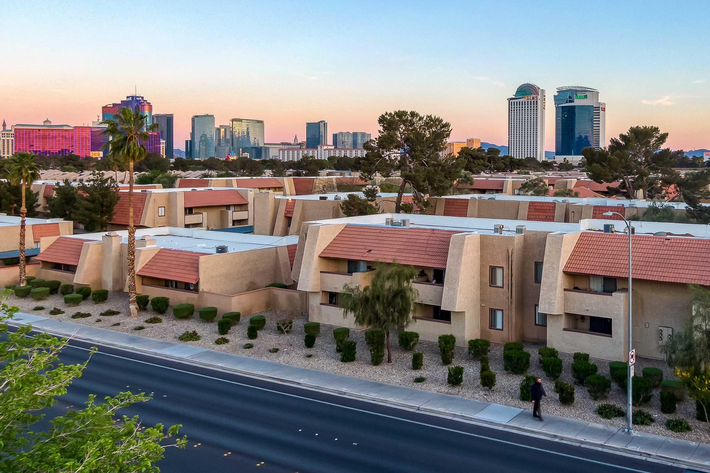 A view of a city skyline at dusk, featuring a mix of modern skyscrapers and residential buildings. In the foreground, low-rise apartments with terracotta roofs are set alongside a road lined with bushes and palm trees, while the sky displays soft pastel colors from the sunset.