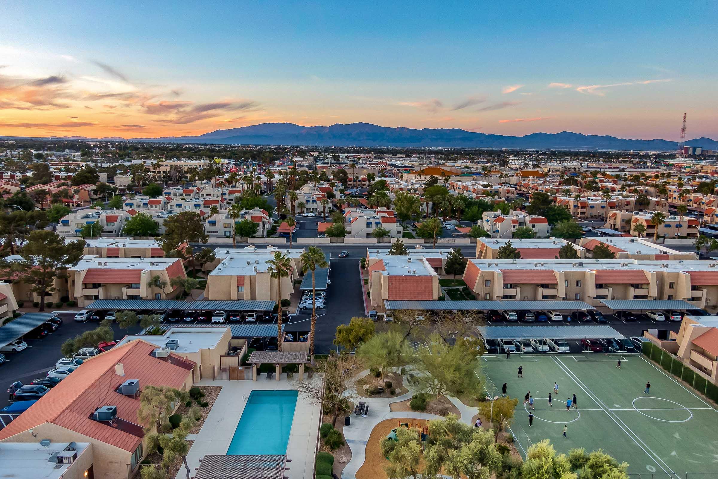 Aerial view of a suburban community featuring houses with red roofs, a swimming pool, and a sports court. The scene is set against a backdrop of mountains and a colorful sunset sky, with parking areas and green spaces visible among the residential buildings.