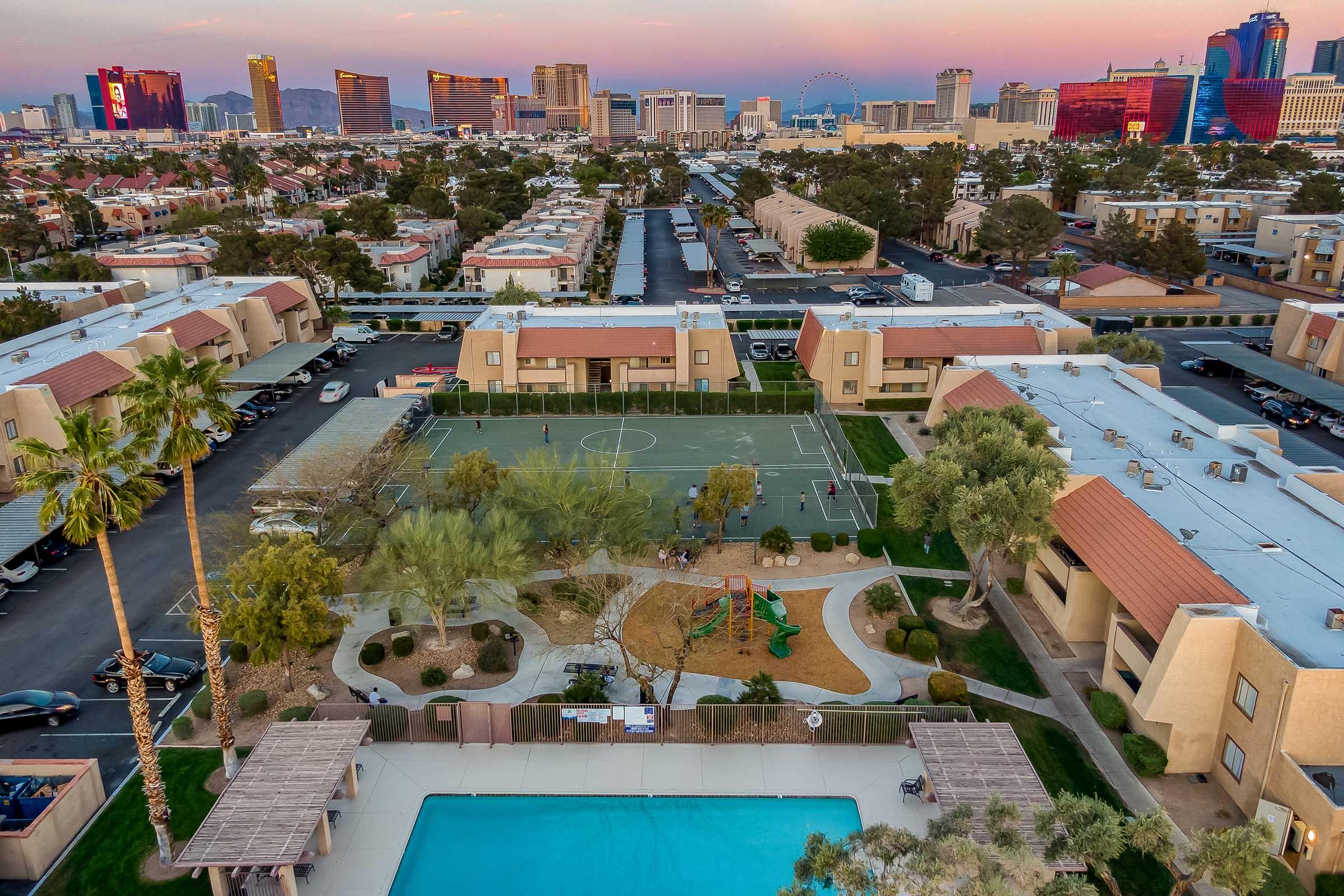 Aerial view of a residential area featuring tennis courts and a playground. In the foreground, a swimming pool is visible, surrounded by landscaped areas. In the background, the skyline of Las Vegas showcases colorful hotels and attractions under a sunset sky.