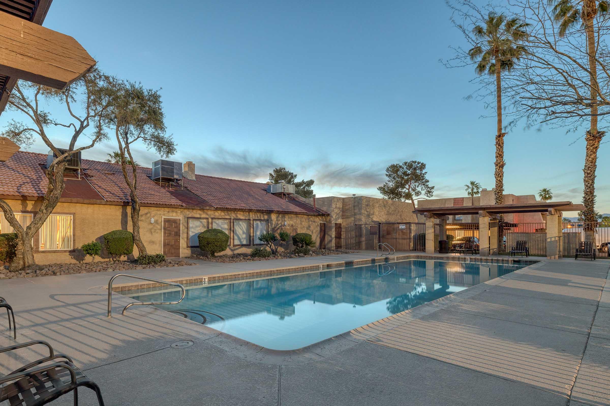 A tranquil pool area featuring a clear blue swimming pool surrounded by lounge chairs. Palm trees and neatly trimmed bushes enhance the serene atmosphere, with a modest building in the background. The scene captures a warm evening ambiance with soft lighting.