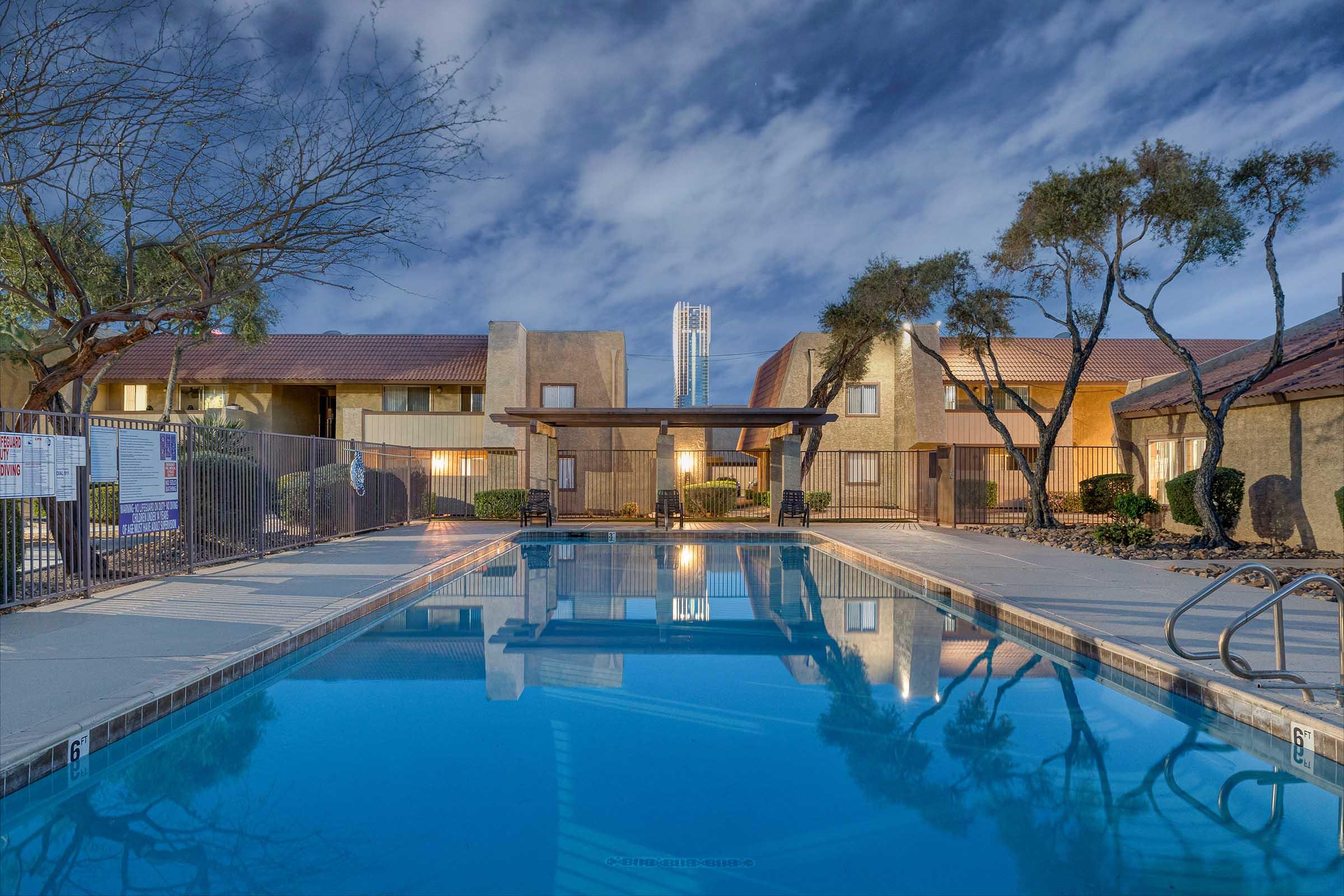 A serene swimming pool surrounded by landscaped areas and apartment buildings. The water reflects the evening sky and nearby trees, with soft lighting creating a calm atmosphere. A few lounge chairs are positioned around the pool, making it an inviting space for relaxation.