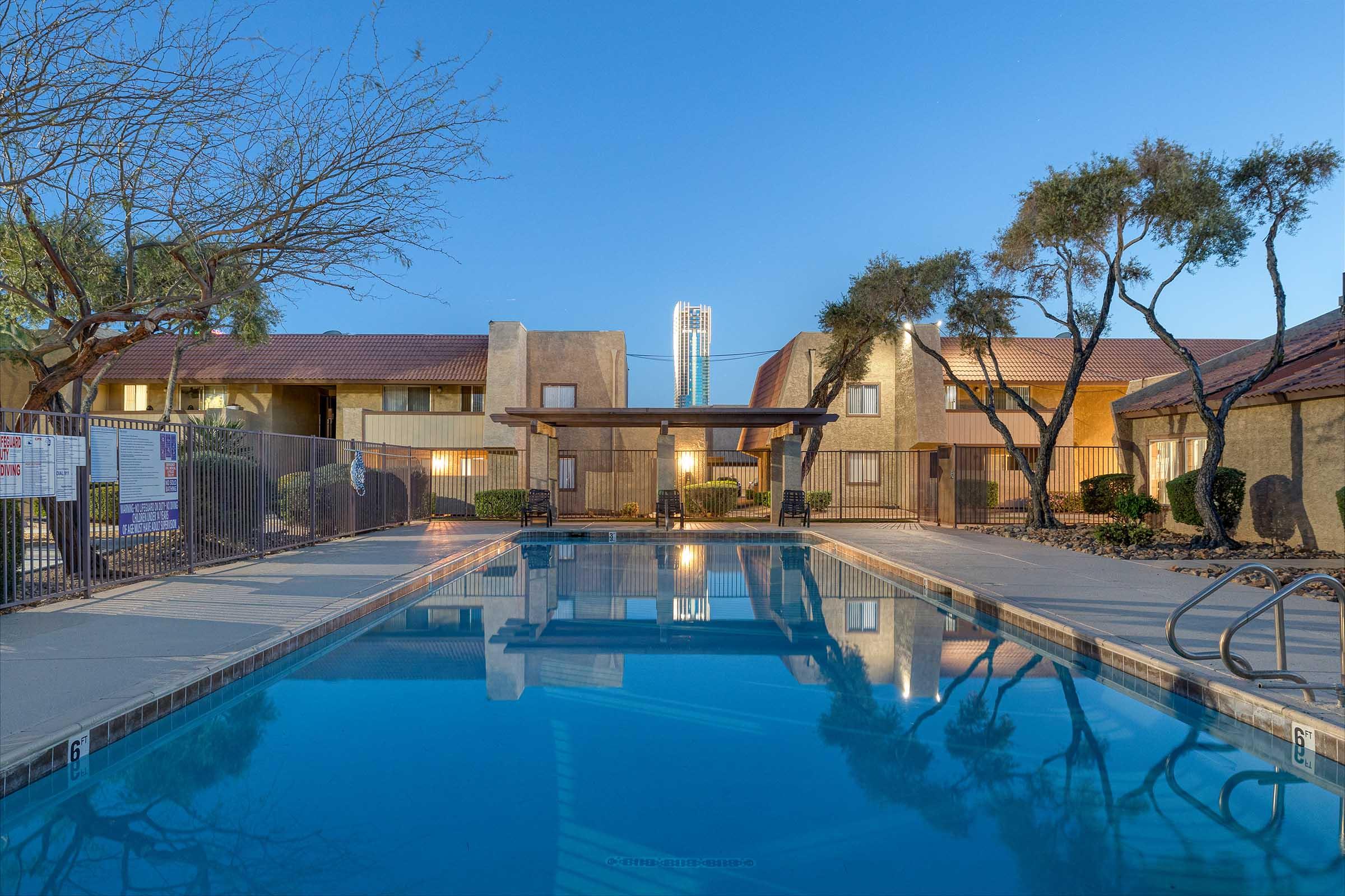 A serene outdoor pool area at dusk, featuring clear, calm water reflecting the surrounding trees and buildings. The setting includes apartment complexes and a pergola, with a warm glow from nearby lights and a clear sky in the background. The atmosphere is tranquil and inviting.