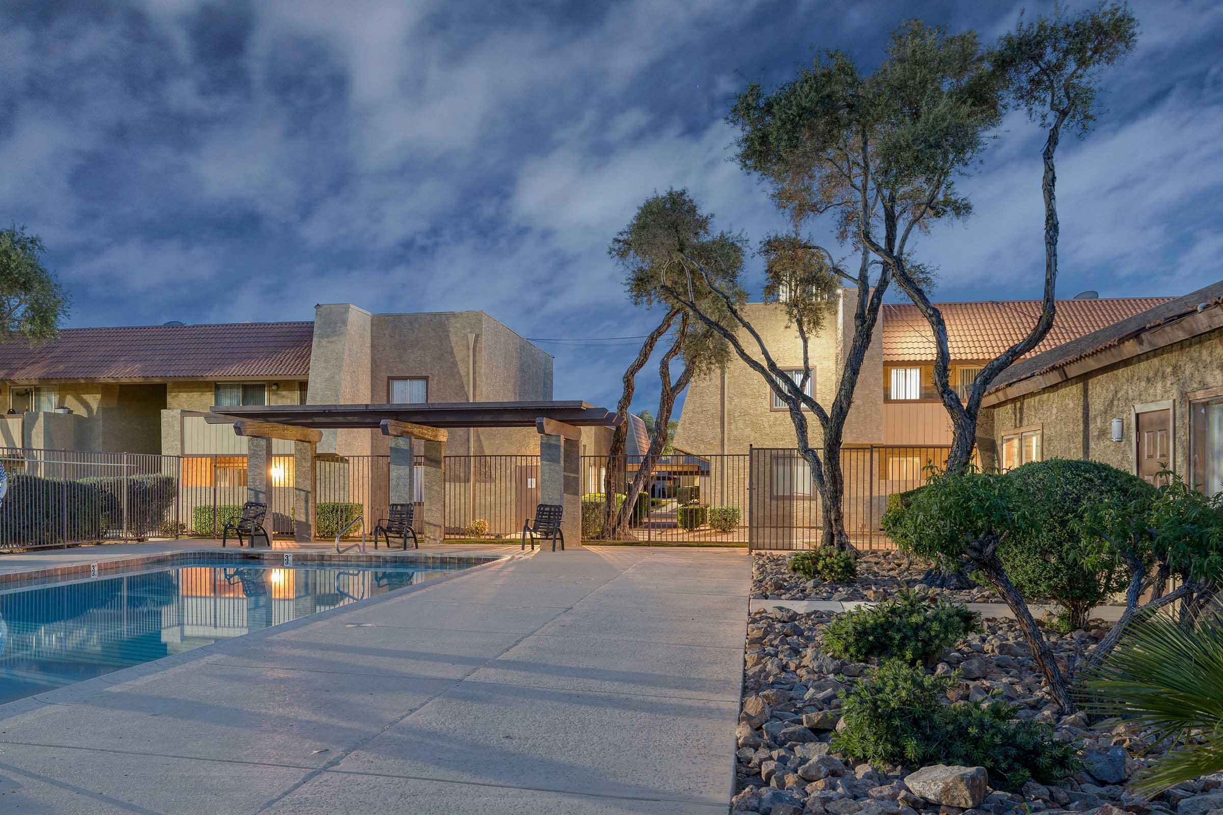 A tranquil pool area surrounded by landscaped trees and shrubs, with lounge chairs set up near the water. Nearby buildings feature a mix of textured walls and tiled roofs, all under a dramatic evening sky filled with clouds. The scene conveys a peaceful atmosphere ideal for relaxation.