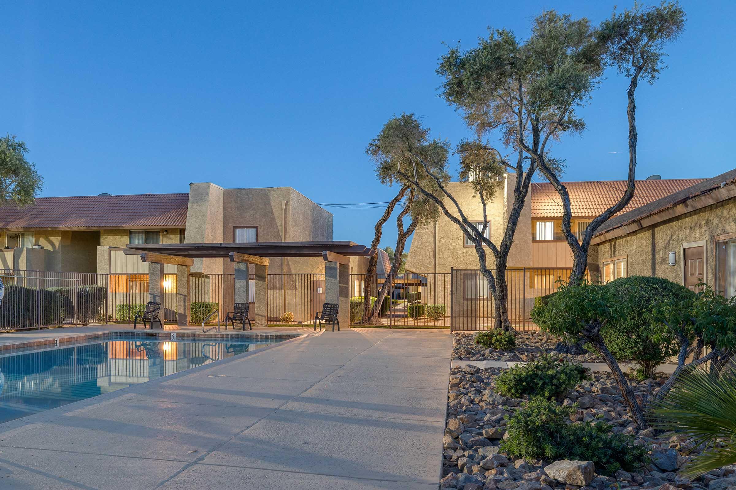 A serene swimming pool area surrounded by landscaped gardens and trees, with a few chairs and a fence. In the background, there are two beige apartment buildings, and the scene is set during twilight, with soft lighting creating a calm atmosphere.