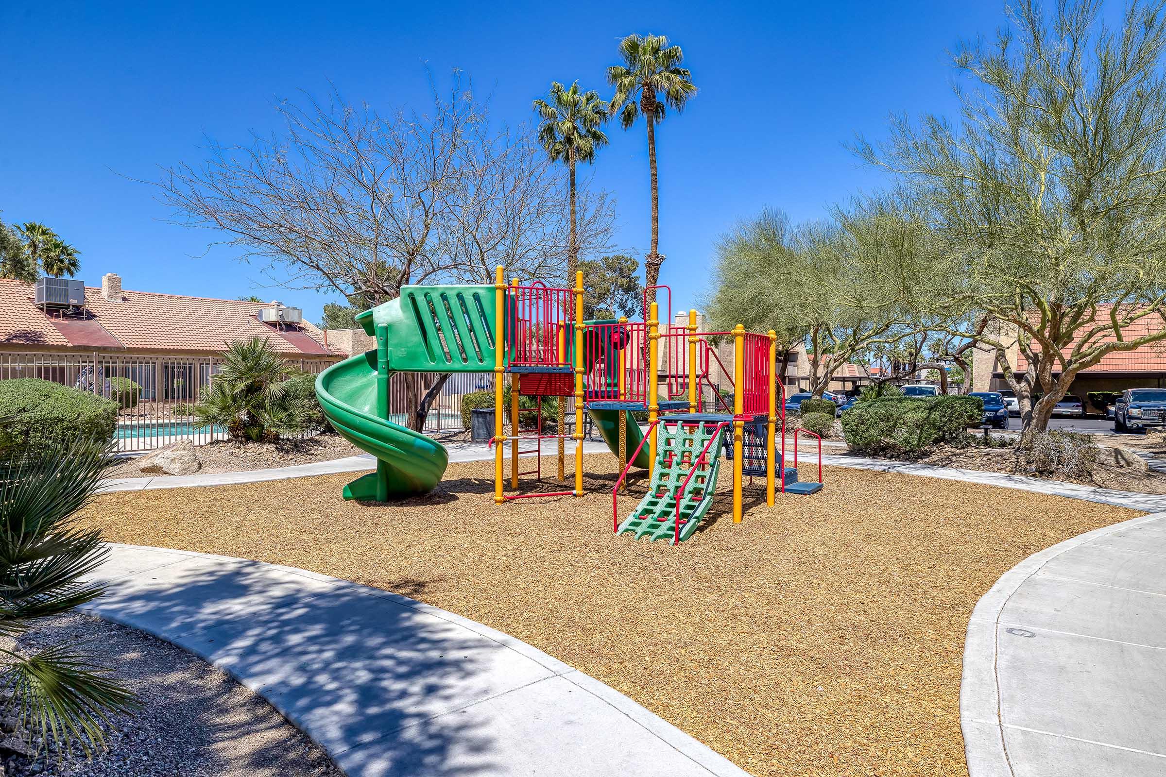 Colorful playground equipment featuring a green slide, climbing structures, and railings, surrounded by gravel and desert landscaping with palm trees under a clear blue sky. Nearby homes are visible in the background, creating a pleasant recreational space.